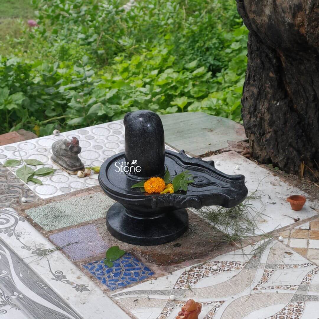 A sacred 12-inch Shivlinga statue exquisitely carved from blackstone is centrally placed on a tiled outdoor surface likely a home temple area surrounded by lush greenery and the trunk of a large tree. The Shivlinga a symbolic representation of Lord Shiva consists of an upright cylindrical pillar emerging from a base signifying the divine creative energy and universal consciousness. The blackstone material gives the statue a profound revered appearance. The 12-inch blackstone Shivlinga is adorned with devotional offerings. A vibrant orange marigold flower and a few fresh green leaves are gently placed on top of the lingam and the yoni base symbolizing devotion and purity. The yoni base itself is intricately shaped featuring a spout to channel sacred offerings poured over the lingam. The dark polished surface of the blackstone gleams subtly reflecting the natural light. Nearby a small terracotta diya and other offerings like betel leaves or fruits are visible indicating an active worship space. A small Nandi figure Shiva's vahana is also partially visible in the background to the left. This blackstone Shivlinga statue ideal for a home temple is perfectly suited for an article discussing Shivlinga statues for home use. The natural setting combined with the traditional offerings creates an authentic and peaceful ambiance for worship. The compact 12-inch size makes it suitable for personal altars while the durable blackstone ensures its longevity and sacred presence allowing devotees to connect with the divine energies of Lord Shiva within their home.