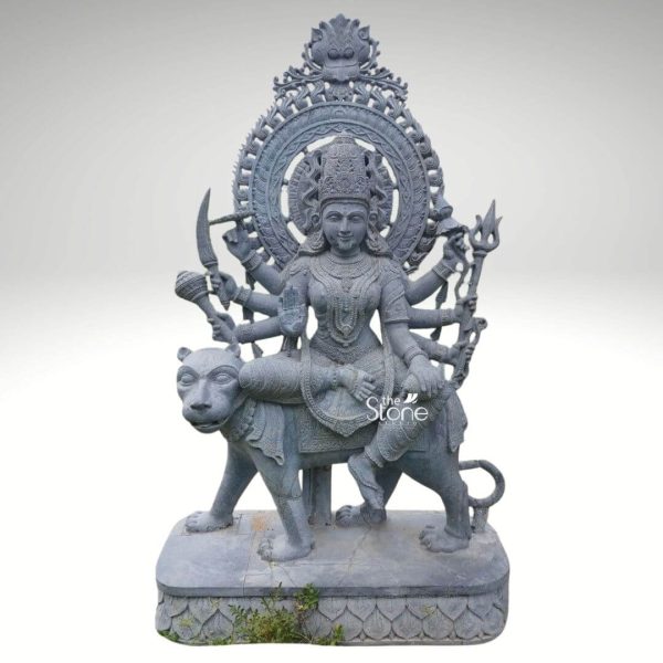 A magnificent 5-foot blackstone sculpture of Durga Maa, ideal for a temple commands attention with its intricate details and powerful presence. The goddess Durga is depicted seated regally on her vahana, a powerful lion both meticulously carved from the dark stone. Durga is adorned with a detailed crown elaborate jewelry including necklaces and armlets and a serene, benevolent expression on her face. Her numerous arms extend gracefully each holding a distinct divine weapon or symbol a sword, a trident with flames, a conch shell and other implements all signifying her protective and destructive powers against evil. Behind her head, a grand and intricately carved circular prabhavali features delicate scrollwork and traditional motifs, culminating in a decorative element at the very top. The lion, her mount, is depicted with a robust body and a watchful gaze conveying strength and loyalty as it strides forward. The entire composition rests on a multi-tiered, carved blackstone base which features subtle decorative patterns. This substantial and beautifully crafted statue embodies the divine energy and grace of Durga making it a perfect focal point for worship and admiration in a temple setting.