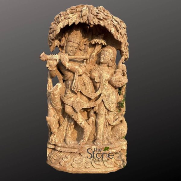 magnificent, intricately carved light-brown sandstone sculpture, depicting the divine couple, Lord Krishna and Radha, under a lush, overarching tree canopy. The vertical sculpture showcases exceptional craftsmanship and traditional Indian artistry. On the left, Lord Krishna stands in his iconic tribhanga (triple-bend) pose, gracefully leaning with one leg crossed, captivatingly playing his bansuri (flute) to his lips. He wears an elaborate crown, possibly adorned with a peacock feather, delicate necklaces, armlets, and anklets, all rendered with exquisite detail. His draped dhoti and flowing sash are meticulously carved, and his serene, blissful expression reflects divine tranquility. Beside him on the right, Radha stands equally poised and elegant, gently turned towards Krishna in a loving gaze. She holds a small, ornate kalash (pot or vessel), possibly symbolizing offerings or nurturing, and is adorned with similar intricate jewelry and gracefully draped garments. Her expression is soft and devoted, complementing Krishna's. Above the figures, the dense, leafy canopy of the tree is deeply carved, forming a natural arch that provides a sense of sacred shelter and a backdrop reminiscent of the groves of Vrindavan. Subtle animal motifs enhance the pastoral scene: a small calf or cow peeks from behind Krishna on the lower left, and a regal peacock is carved into the foliage on Radha's right, its feathers detailed. The entire composition rests on a robust, multi-tiered pedestal, meticulously carved with traditional Indian patterns and scrollwork, providing a solid and ornate foundation for the divine narrative. The logo "The Stone Studio" is subtly carved into the bottom front of the base. This piece exemplifies exquisite Hindu devotional art, showcasing the beauty of sandstone carving and its connection to spiritual traditions and cultural heritage.
