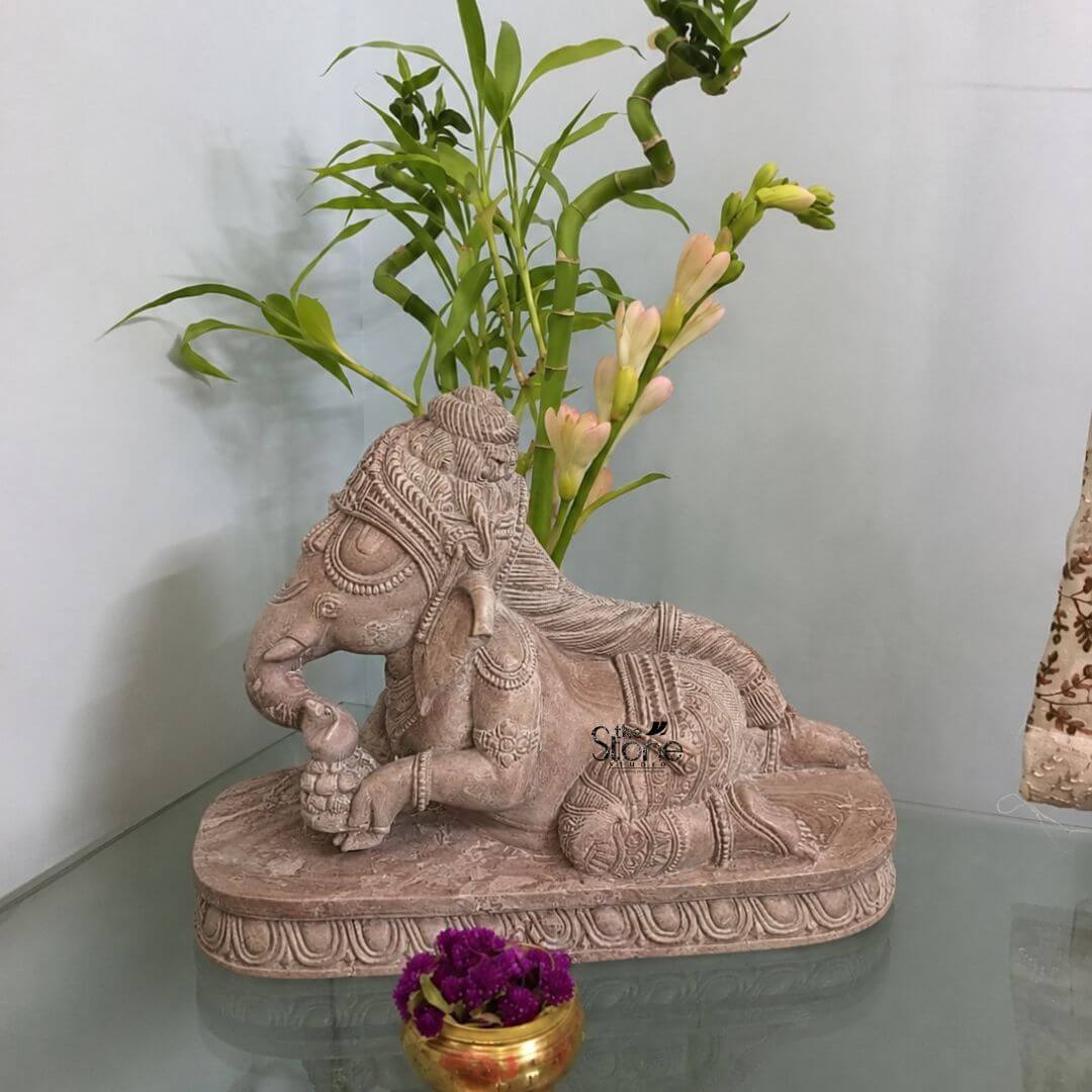Crawling Ganesha Statue in Pink Stone 1ft 3 A detailed medium shot captures a beautiful one-foot-tall statue of Baby Ganesha hand-carved from a warm pink sandstone. The statue perfectly sized for home decor is depicted in a charming playful crawling posture. Lord Ganesha is resting on a matching ornately carved rectangular pedestal with a traditional lotus petal border. His large elephant head features a serene joyful expression with intricate detailing on the ornate crown and large expressive ears. He is wearing delicate carved jewelry including a rudraksha mala and armlets with his trunk curled and holding a small sweet or laddu. The pink stone sculpture exhibits a rich texture and detailed craftsmanship throughout its form. The deity's body shows realistic folds of skin and traditional cloth all rendered in the soft natural hue of the sandstone. The statue's surface is matte and slightly rough to the touch emphasizing the hand-carved quality. The Ganesha figure is positioned centered on a reflective glass surface likely a shelf or tabletop which subtly mirrors the statue and pedestal. The background provides a tasteful uncluttered setting for this home decor piece. A light neutral-toned wall creates a soft backdrop with a partial view of patterned, natural-fiber curtains to the right. Directly behind the statue a live lucky bamboo plant with several twisted stalks adds a touch of vibrant green and natural life. In the foreground placed on the glass surface just in front of the pedestal is a small gleaming brass pot filled with a cluster of deep purple Gomphrena flowers adding a rich color accent that complements the pink stone. The overall composition is balanced and serene highlighting the statue as a focal point for interior decoration.