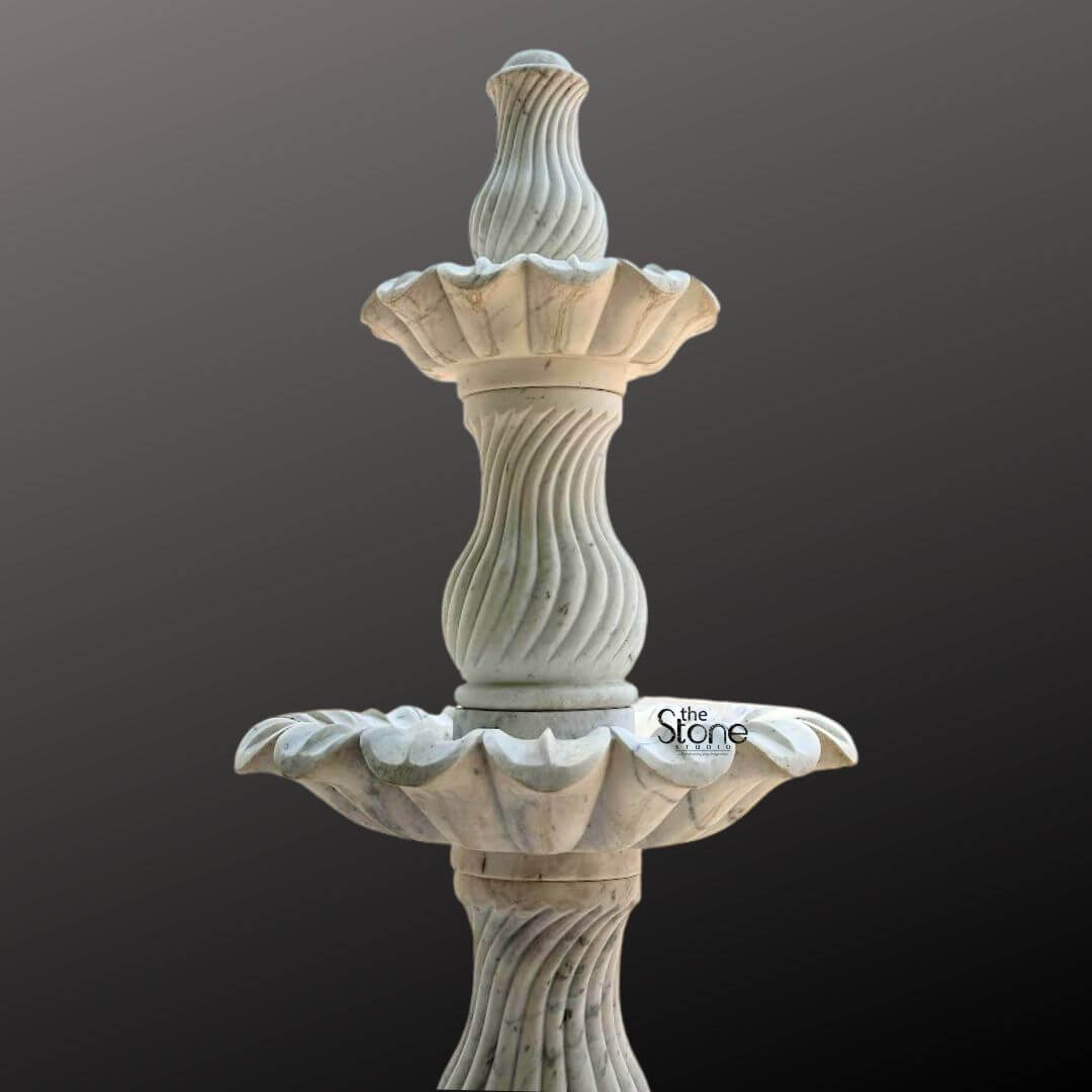 Marble Fountain Sculpture 6ft 5 A grand 6-foot tall three-tiered marble fountain sculpture is prominently displayed against a plain dark gray background showcasing its elaborate design and impressive scale suitable for garden decor. The fountain is expertly carved from light-colored marble exhibiting a natural white or off-white hue with subtle gray veining and a smooth polished finish. The sculpture features three distinct levels of basins each progressively larger towards the bottom designed to create a magnificent cascading water effect. The uppermost tier consists of a small fluted basin crowned with an elegant finial where water would typically originate. Below this the second tier expands into a wider beautifully scalloped basin designed to catch and spill water. The largest and lowest visible basin is also exquisitely scalloped with a pronounced floral or shell-like edge that adds to its ornamental appeal. Connecting these basins are intricately carved central pillars. The upper two visible pillars feature elegant spiral fluting adding a dynamic and classical visual element while the lowest visible pillar continues the sophisticated fluted design providing a strong and artistic base. The entire fountain rests on a solid round pedestal that transitions smoothly into its base though the very bottom is slightly cropped. The meticulous craftsmanship highlights traditional European or classical garden aesthetics making this marble fountain a timeless and luxurious addition to a large garden, courtyard or park, promising both visual splendor and the soothing sound of flowing water perfectly suited to enhance any outdoor space.