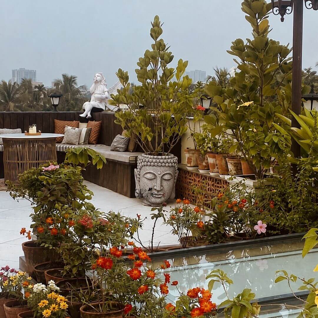 A lush and inviting terrace garden is depicted featuring a central focal point of a beautifully crafted Buddha Pot Statue for Terrace Garden. The statue which stands approximately 3 feet tall is made of durable grey fibreglass giving it the appearance of carved stone while remaining lightweight. It is sculpted in the serene likeness of a Buddha head, with downcast eyes a calm expression and a detailed Ushnisha. The top of the head is designed as a deep planter currently holding a vibrant leafy green plant with broad, rounded leaves seamlessly integrating the spiritual element with the garden's greenery. The 3-foot Buddha Fibre Planter statue is surrounded by an abundance of potted plants creating a verdant and tranquil oasis on the rooftop. In the foreground a variety of terracotta pots hold colorful flowers including bright orange and red blooms as well as yellow daisies adding depth and a riot of color. To the left a built-in wooden bench with patterned cushions offers a comfortable seating area adjacent to a small rustic rattan table. Behind the Buddha planter, taller plants in terracotta pots line a decorative wooden screen further enhancing the natural and private feel of the space. This image perfectly illustrates the article's theme of incorporating meaningful decor into a rooftop space by showcasing the Buddha head planter as a central element. The substantial scale of the 3-foot statue makes it a commanding presence that harmonizes with the surrounding foliage demonstrating its ability to transform a simple terrace into a peaceful sanctuary. The scene is set against a backdrop of distant high-rise buildings and palm trees under an overcast sky anchoring the urban garden in its metropolitan context and highlighting the potential for creating a personal retreat in a city.