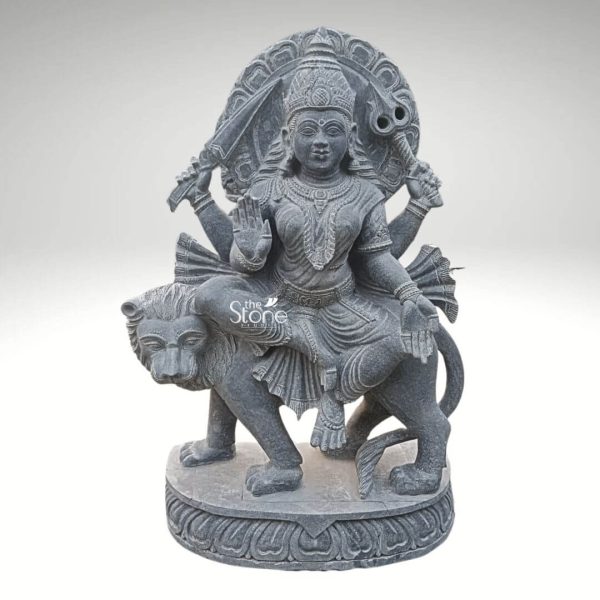 A powerful 33-inch Durga Maa statue carved from dark black stone depicts the goddess seated majestically upon her lion mount ideal for an article about Durga Maa Lion Statue. Durga Maa is intricately adorned with a crown necklaces armlets and other jewelry all finely detailed in the stone. Her expression is serene yet commanding signifying her divine authority. She is depicted with multiple arms each holding various divine weapons and symbols. Prominently she holds a sword in one hand while another hand is raised in a gesture of blessing or fearlessness . Behind her head a grand, circular prabhavali is elaborately carved with traditional patterns and a decorative element at the top enhancing her divine aura. The lion, Durga's vahana is portrayed with strength and vitality, its mane detailed and its posture dynamic as it strides forward. The entire sculpture rests on a sturdy round black stone base, which is also adorned with lotus petal carvings and other traditional motifs providing a solid foundation and enhancing its aesthetic appeal. The dark black stone emphasizes the intricate carving and the powerful presence of both Durga and her lion.
