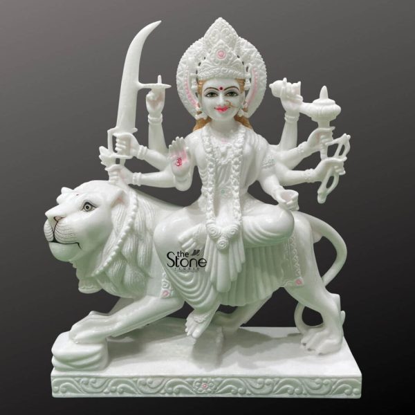 This image showcases a stunning 2-foot Durga Maa statue meticulously carved from pristine white marble depicting the goddess seated powerfully on her lion mount. The statue is presented against a dark contrasting background which makes the pure white marble glow and highlights every intricate detail. Durga Maa is depicted with a serene yet determined expression her face featuring delicate painted details like red lips defined eyebrows and a red bindi bringing her divine presence to life. She is adorned with an elaborate crown and traditional jewelry including necklaces earrings, and bangles, all rendered in exquisite detail within the marble. The goddess possesses eight arms, each gracefully holding a distinct weapon or symbolic item representing her various divine powers. These include a sword a trident, a conch shell and a discus among others. Her primary right hand is raised in a gesture of fearlessness or blessing while her left hand cradles an item. She is draped in finely sculpted marble drapery that flows elegantly conveying movement and grace even in stone. Durga Maa's vehicle a majestic lion is equally detailed and powerful. The lion is sculpted in a dynamic stance with a flowing mane muscular body and alert expression symbolizing strength courage and protection. The goddess sits comfortably on the lion's back a testament to her command over creation. Both figures are mounted on a rectangular multi-tiered white marble base which features subtle decorative carvings along its edges. This 2 feet Durga marble statue sitting on her vahan is a magnificent representation of divine power and beauty, perfectly suited for an article discussing such an idol.