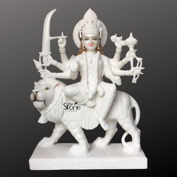 This impressive image showcases a 2.5-foot Durga Maa statue meticulously carved from pristine white marble depicting the goddess seated powerfully on her majestic tiger mount. The statue is presented against a dark contrasting background which makes the pure white marble glow and highlights every intricate detail making it a focal point for an article on Durga Maa with Tiger Statue in Marble. Durga Maa is rendered with a serene yet determined expression her face featuring delicate painted details such as red lips defined eyebrows and a red bindi bringing a vibrant lifelike quality to her divine presence. She is adorned with an elaborate crown and traditional jewelry, including necklaces earrings and bangles all exquisitely sculpted into the marble. The goddess is depicted with eight arms each gracefully holding a distinct weapon or symbolic item representing her various divine powers. These include a prominent sword held high, a trident a conch shell, a bow and arrows, among others. Her primary right hand is raised in a gesture of fearlessness or blessing, while her left hand cradles an item. She is draped in finely sculpted marble drapery that flows elegantly, conveying movement and grace even in stone. Durga Maa's vehicle a powerful tiger is equally detailed and commanding. The tiger is sculpted in a dynamic stance, with a richly textured mane muscular body, and an alert formidable expression symbolizing strength, courage and protection against evil. The goddess sits comfortably on the tiger's back, a testament to her supreme command. Both figures are mounted on a solid, rectangular, multi-tiered white marble base which features subtle decorative carvings along its edges. This 2.5 feet Durga marble statue is a magnificent representation of divine power, beauty, and protection, perfectly suited for an article discussing such an idol.