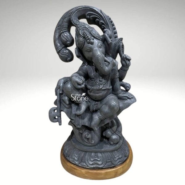 An exquisitely hand-carved 24-inch Ganesha murti in black stone masterfully sculpted from premium natural black stone to serve as a majestic spiritual centerpiece for a home or office. This 2-foot tall sculpture captures Lord Ganesha in a stable and serene seated posture atop an intricately detailed circular lotus pedestal radiating an aura of peace and divine stability. Lord Ganesha is portrayed with his iconic elephant head featuring a towering tiered crown large expressive ears that represent the wisdom of listening, and a gracefully curved trunk. The deity is shown with four arms holding sacred spiritual symbols, including traditional implements and a bowl of modaks representing the sweetness of spiritual fulfillment. The craftsmanship highlights incredible artisan detail in the layered necklaces, armlets, and realistic fabric folds of his traditional attire. Framing the deity is a grand, circular Prabhavali embellished with rhythmic floral and scrollwork patterns that symbolize a divine aura. The refined matte charcoal finish and grainy texture of the natural stone highlight the heritage of traditional Indian stone masonry making this black stone Ganesha statue a perfect addition to a home mandir or entrance to invite prosperity, protection, and the removal of obstacles.