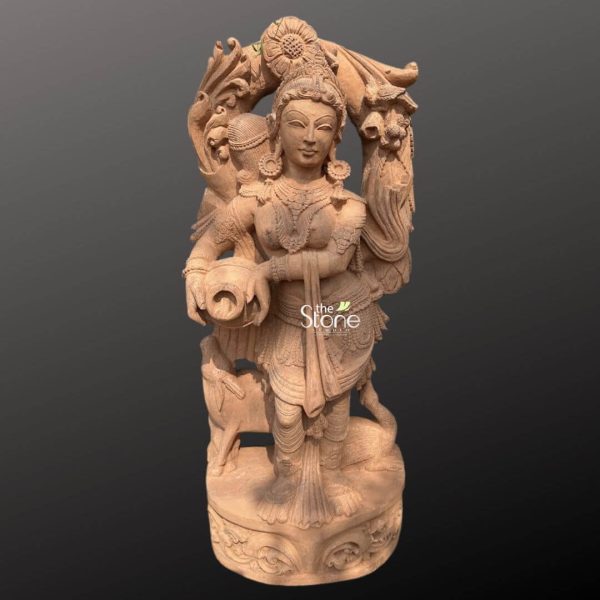 An exquisitely hand-carved 4-foot tall sandstone Devadasi statue masterfully designed as a graceful garden angel centerpiece for a premium landscape. This spiritual sculpture crafted from natural brown sandstone depicts a traditional celestial maiden in a serene standing posture gracefully holding a water pot under one arm. The figure is adorned with incredibly detailed traditional jewelry including layered necklaces ornate waist belts, and large earrings, showcasing the depth of expert stone masonry. Lord Devadasi is framed by a grand circular backplate featuring rhythmic floral relief carvings and a sunflower motif at the peak. At the base of the sculpture small lifelike figures of a cow and a swan are subtly carved, symbolizing innocence, purity, and devotion. The grainy, earthy texture and rustic finish of the natural sandstone provide a timeless antique aesthetic that blends perfectly with outdoor greenery. This durable and weather-resistant large-scale stone sculpture serves as an ideal focal point for home gardens, courtyards, or outdoor sanctuaries, inviting an atmosphere of peace, elegance, and divine protection into the environment.
