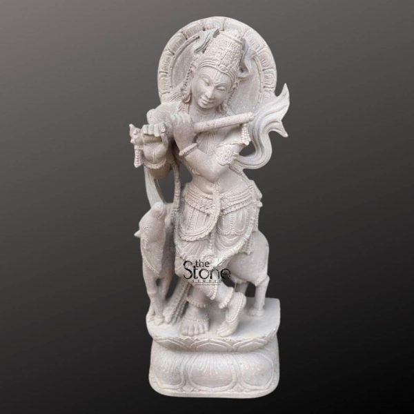 A beautifully carved 2.5-foot Krishna sculpture fashioned from light-colored Rajasthani sandstone is prominently displayed against a deep dark grey background highlighting its intricate details. Lord Krishna is depicted standing in a graceful Tribhanga pose playing his divine flute which he holds to his lips. The sculpture showcases meticulous craftsmanship typical of Rajasthani stone artistry. Krishna is adorned with an elaborate crown intricate jewelry including necklaces armlets, and anklets and flowing garments all finely rendered in the sandstone. His facial features are serene and benevolent, with a gentle smile and expressive eyes that convey a sense of divine charm. Behind his head an ornate circular halo or aureole with radiating patterns is carved symbolizing his divine presence. Further enhancing the pastoral scene, a sacred cow or calf is sculpted standing gently beside Krishna on his right looking upwards towards him. On his left a smaller elephant figure is depicted, adding to the rich narrative. The entire 2.5-foot Krishna statue stands on a multi-tiered lotus pedestal also carved from sandstone providing a stable and traditional base. The light earthy tone and subtle texture of the Rajasthani sandstone give the sculpture a natural warm and inviting aesthetic. This detailed Krishna sculpture with flute in Rajasthani stone beautifully embodies love, joy, and spiritual harmony, directly aligning with an article discussing such revered idols.