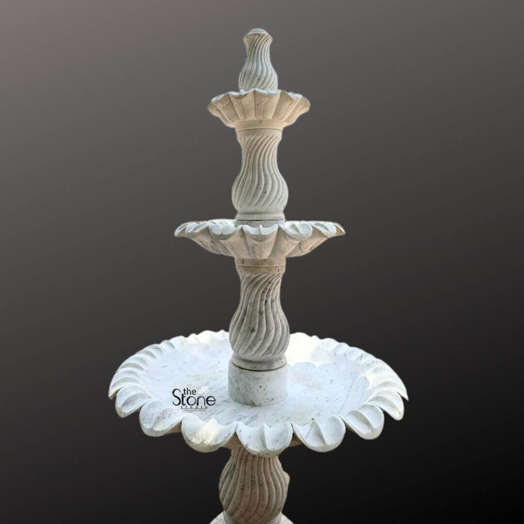 Marble Fountain Sculpture 6ft 2 A grand 6-foot tall three-tiered layer fountain expertly carved from marble is prominently displayed against a plain dark gray background showcasing its intricate design and suitability for garden ornamentation. The fountain is crafted from light-colored marble exhibiting a natural white or off-white hue with subtle gray veining and a smooth polished finish. The sculpture features three distinct levels of basins each progressively larger towards the bottom designed to create a magnificent cascading water effect. The uppermost tier consists of a small fluted basin crowned with an elegant finial where water would typically originate. Below this the second tier expands into a wider beautifully scalloped basin designed to catch and spill water. The largest and lowest visible basin is also exquisitely scalloped with a pronounced floral or shell-like edge that adds to its ornamental appeal. Connecting these basins are intricately carved central pillars. The upper two visible pillars feature elegant spiral fluting adding a dynamic and classical visual element while the lowest visible pillar continues the sophisticated fluted design providing a strong and artistic base. The entire fountain rests on a solid round pedestal that transitions smoothly into its base. The meticulous craftsmanship highlights traditional European or classical garden aesthetics, making this marble layer fountain a timeless and luxurious addition to a large garden courtyard, or park promising both visual splendor and the soothing sound of flowing water perfectly suited to enhance any outdoor space.