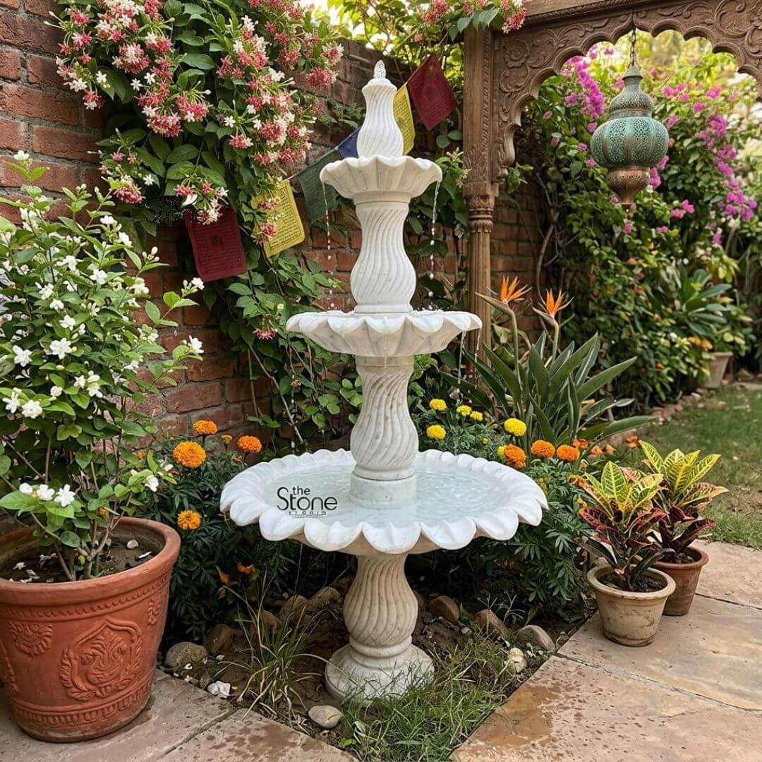Marble Fountain Sculpture 6ft 3 A stunning 6-foot tall three-tiered layer fountain exquisitely carved from white marble serves as a magnificent centerpiece in a lush and vibrant garden landscape. The fountain is expertly crafted showcasing a natural white hue with subtle variations and its smooth polished surface reflects the surrounding light. The fountain features three distinct levels of basins each progressively larger towards the bottom designed to create a beautiful cascading water effect. The uppermost tier is a small, fluted basin topped with an elegant finial from which water gently trickles. Below it the second tier features a wider beautifully scalloped basin catching the falling water. The largest and lowest basin is also exquisitely scalloped with a pronounced floral or shell-like edge that adds to its ornamental appeal and its surface is visibly filled with water. Connecting these basins are intricately carved central pillars. The upper two pillars feature elegant spiral fluting adding a dynamic and classical visual element while the lowest visible pillar continues the sophisticated fluted design providing a strong and artistic base. The fountain rests on a solid round pedestal that blends seamlessly with the surrounding garden bed. The garden environment is rich and diverse featuring a rustic brick wall covered in climbing flowering plants with pink and white blossoms. Small prayer flags hang against the brick wall. Various green plants including potted foliage with variegated leaves and beds of orange and yellow marigold-like flowers adorn the foreground. In the background a decorative wooden archway and a traditional hanging lantern with a greenish hue add architectural and cultural interest. This marble layer fountain is a timeless and luxurious addition to any landscape providing both visual splendor and the soothing sound of flowing water perfectly enhancing the natural beauty and tranquility of the garden.