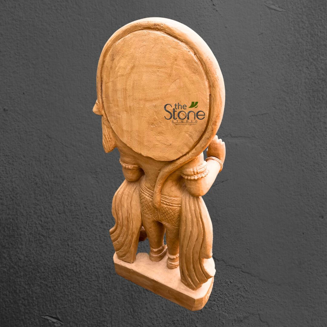 Hanuman Statue in Red Stone Standing Position 3ft 2 beautifully carved, natural-toned statue, likely made of wood or sand-colored stone, is presented from a three-quarter rear view against a dark, textured gray background. The craftsmanship is highly detailed, showcasing a standing figure with a slender form. A prominent, smooth, and perfectly circular halo or aureole is positioned behind the figure's head, which is visible in profile looking towards the left. The back of the figure's torso is visible, leading down to a waist adorned with a patterned band or belt. Flowing drapery gracefully cascades around the lower body and legs, exhibiting intricate folds and a delicate, textured pattern that suggests a traditional garment like a dhoti. The arms are bent at the elbows, with hands positioned near the chest, though the specific gestures (mudras) are not fully discernible from this angle. Glimpses of ornamental bangles or bracelets can be seen on the wrists, adding to the elegance of the piece. The entire statue rests on a simple, sturdy rectangular base, carved from the same material. The soft lighting highlights the texture and contours of the carving, emphasizing the artisan's skill. A "the Stone Studio" logo, featuring a stylistic leaf motif, is subtly watermarked on the upper right side of the halo. This image vividly represents the studio's expertise in creating traditional Indian or Hindu sculptures, perhaps depicting a deity, mythological figure, or celestial being, making it a perfect visual to accompany an article discussing spiritual art, cultural craftsmanship, or unique home decor pieces.