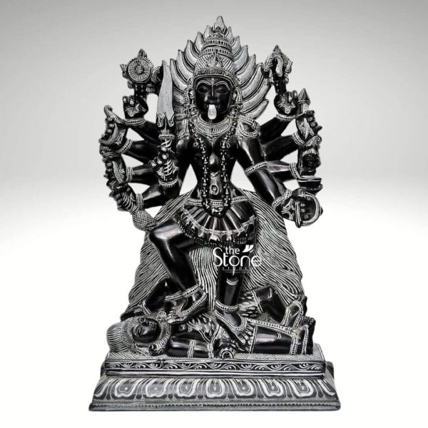 A powerful 1-foot Maa Kali statue carved from black stone is dramatically presented against a plain light grey background. The goddess Kali is depicted in her fierce and dynamic form standing triumphantly with her right foot placed firmly on the chest of Lord Shiva who lies supine beneath her. This iconic pose often referred to as Kali standing on Shiva symbolizes the triumph over ego and the ultimate reality where even Shiva bows to her cosmic dance of destruction and creation. Maa Kali is intricately sculpted with numerous arms each wielding a different weapon or symbolic item emphasizing her omnipotence and ability to conquer evil. Her face though dark conveys a fierce expression with a protruding tongue typical of her iconography. She is adorned with a garland of skulls a skirt of severed arms and an elaborate crown all meticulously detailed in the black stone. The fine carving captures the texture of her hair the details of her ornaments and the powerful musculature implied beneath her dark skin. Lord Shiva lying beneath her is also detailed depicted in a state of calm surrender. The entire scene rests on a multi-tiered black stone base which is decorated with traditional motifs adding to the grandeur and spiritual significance of the sculpture. The deep black color of the stone enhances the dramatic impact and sacredness of the scene. This detailed 1-foot Maa Kali statue standing on Shiva is a profound representation of divine power and liberation perfectly aligning with an article discussing such an iconic black stone sculpture.
