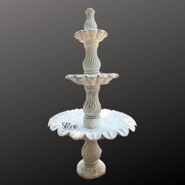 A stately 6-foot tall three-tiered marble fountain sculpture is prominently displayed against a plain dark gray background showcasing its intricate design and potential as a grand garden feature. The fountain is expertly carved from light-colored marble exhibiting a natural white or off-white hue with subtle gray veining and a smooth polished finish. The sculpture features three distinct levels of basins each progressively larger towards the bottom designed to create a cascading water effect. The top tier is a small fluted basin topped with a finial from which water would typically emerge. Below it the second tier features a wider scalloped basin. The largest and lowest basin is also beautifully scalloped with a pronounced floral or shell-like edge that catches the eye. Connecting these basins are intricately carved central pillars. The upper two pillars feature elegant spiral fluting adding a dynamic visual element while the lowest pillar is wider and also has a subtle fluted design providing a strong base. The entire fountain rests on a solid round pedestal that transitions smoothly into its base. The craftsmanship highlights traditional European or classical garden aesthetics making this marble fountain a timeless and elegant addition to a large garden courtyard, or park promising both visual beauty and the soothing sound of flowing water.