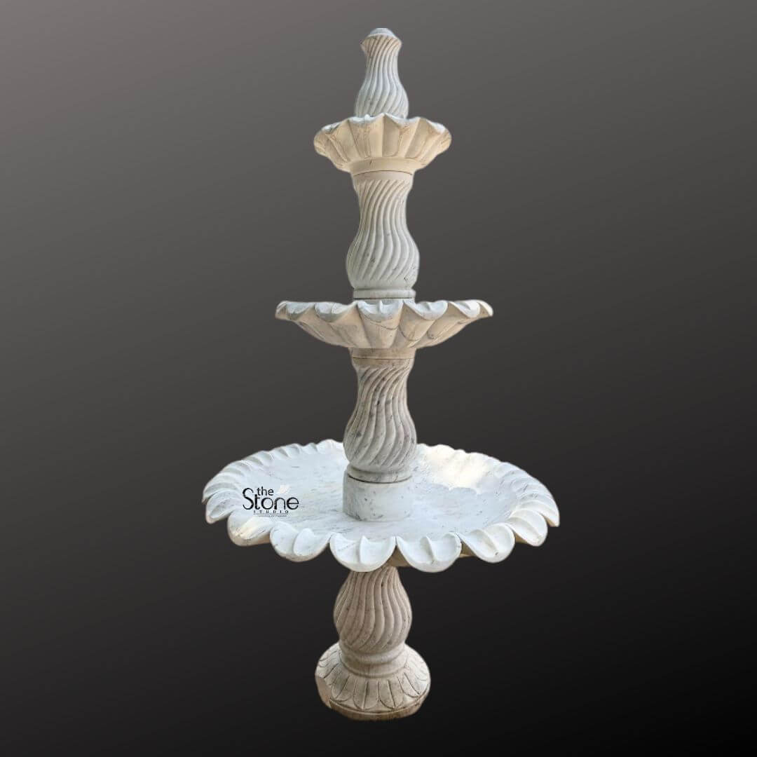 Marble Fountain Sculpture 6ft 4 A grand 6-foot tall three-tiered marble fountain statue is prominently displayed against a plain dark gray background showcasing its intricate design and impressive scale ideal for a garden setting. The fountain is expertly carved from light-colored marble exhibiting a natural white or off-white hue with subtle gray veining and a smooth polished finish. The sculpture features three distinct levels of basins each progressively larger towards the bottom meticulously designed to create a magnificent cascading water effect. The uppermost tier consists of a small fluted basin crowned with an elegant finial where water would typically originate. Below this the second tier expands into a wider beautifully scalloped basin designed to catch and spill water. The largest and lowest visible basin is also exquisitely scalloped with a pronounced floral or shell-like edge that adds to its ornamental appeal. Connecting these basins are intricately carved central pillars. The upper two visible pillars feature elegant spiral fluting adding a dynamic and classical visual element while the lowest visible pillar continues the sophisticated fluted design providing a strong and artistic base. The entire fountain rests on a solid round pedestal that transitions smoothly into its base. The meticulous craftsmanship highlights traditional European or classical garden aesthetics making this marble fountain a timeless and luxurious addition to a large garden, courtyard or park, promising both visual splendor and the soothing sound of flowing water perfectly suited to enhance any outdoor space.