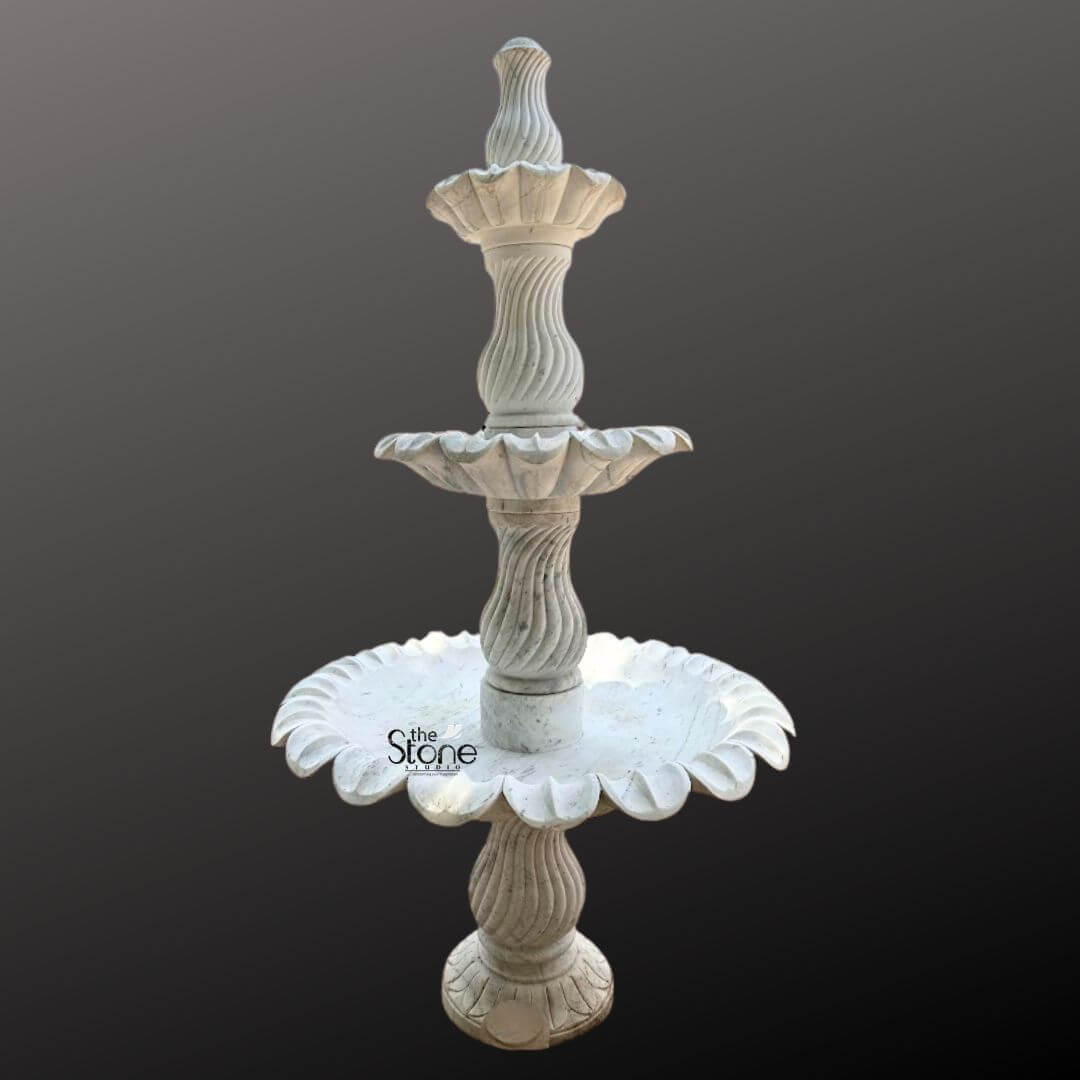 Marble Fountain Sculpture 6ft 6 A grand 6-foot tall three-tiered marble layer fountain is prominently displayed against a plain dark gray background showcasing its intricate design and impressive scale suitable as a luxurious feature for a large home or garden. The fountain is expertly carved from light-colored marble exhibiting a natural white or off-white hue with subtle gray veining and a smooth polished finish. The sculpture features three distinct levels of basins each progressively larger towards the bottom meticulously designed to create a magnificent cascading water effect. The uppermost tier consists of a small fluted basin crowned with an elegant finial where water would typically originate. Below this the second tier expands into a wider beautifully scalloped basin, designed to catch and spill water. The largest and lowest visible basin is also exquisitely scalloped with a pronounced floral or shell-like edge that adds to its ornamental appeal. Connecting these basins are intricately carved central pillars. The upper two visible pillars feature elegant spiral fluting adding a dynamic and classical visual element while the lowest visible pillar continues the sophisticated fluted design providing a strong and artistic base. The entire fountain rests on a solid round pedestal that transitions smoothly into its base. The meticulous craftsmanship highlights traditional European or classical garden aesthetics making this marble layer fountain a timeless and elegant addition to a large home's exterior courtyard or garden promising both visual splendor and the soothing sound of flowing water.
