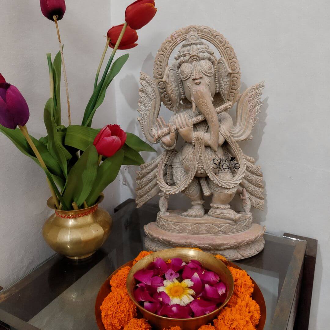 Ganesh Statue Standing with Flute 1ft 2 A detailed medium shot captures a beautiful one-foot-tall statue of the Hindu deity Ganesha hand-carved from a warm pink sandstone making it an exquisite piece for tabletop decor. The statue depicts Ganesha in a graceful standing tribhanga posture holding a flute to his trunk with two hands creating a charming Musical Ganesha representation. He is four-armed adorned with intricately carved traditional jewelry a towering tiered crown and flowing garments all rendered in the delicate pink hue of the stone with exceptional detail. The stone sculpture is placed centrally on a reflective glass-topped side table which provides a modern contrast to the traditional craftsmanship. To the left of the idol sits a traditional polished brass kalash or pot holding a vibrant arrangement of three tulips—one deep purple and two red—providing a striking natural color complement to the monochrome statue. In the foreground centered on the glass surface a circular brass urli bowl is filled with water and a floating floral arrangement of pink rose petals and a central white and yellow chrysanthemum adding a soft welcoming aroma and color to the display. The background is a simple neutral-toned wall ensuring the focus remains entirely on the Ganesha idol and its surrounding decor elements. The lighting is soft and ambient highlighting the rich texture of the hand-carved stone and the warm sheen of the brass vessels. This composition creates a harmonious and inviting arrangement illustrating its suitability as a decorative centerpiece for living spaces or a home temple corner.