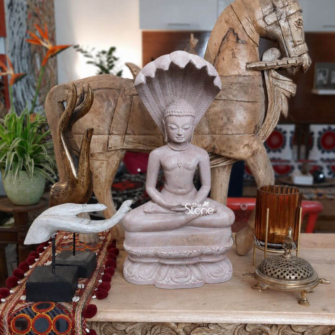 Naga Buddha Statue in Pink Stone 15" 3 A striking 15-inch Naga Buddha Statue meticulously carved from soapstone is prominently displayed on an ornate wooden console table serving as a focal point for home decor. The Buddha is depicted in a meditative seated posture with his hands resting gently in his lap emanating profound peace. Behind his head a multi-headed cobra rises with its hoods spread wide forming a protective canopy which is the defining characteristic of this Naga Buddha form. The soapstone has a soft light natural hue. The soapstone Naga Buddha statue features exquisite details from the serene expression on the Buddha's face with downcast eyes and a subtle smile to the intricate scales and heads of the protecting cobra. His hair is sculpted in tight curls and his robes are draped elegantly across his body revealing some bare skin on his chest. The statue rests on a beautifully carved lotus base adding a traditional touch. In the foreground decorative elements like a carved wooden hand a patterned fabric runner with mirrors and an ornate brass incense burner further enhance the aesthetic. This 15-inch soapstone Naga Buddha statue is an ideal subject for an article on Buddha statues for home decor particularly those with unique symbolic features. In the background a large rustic carved wooden horse figure adds a touch of bohemian elegance while a bird of paradise flower provides a pop of natural color. The entire arrangement creates a rich culturally inspired and peaceful ambiance perfect for bringing a sense of spiritual calm and artistic beauty to any living space.