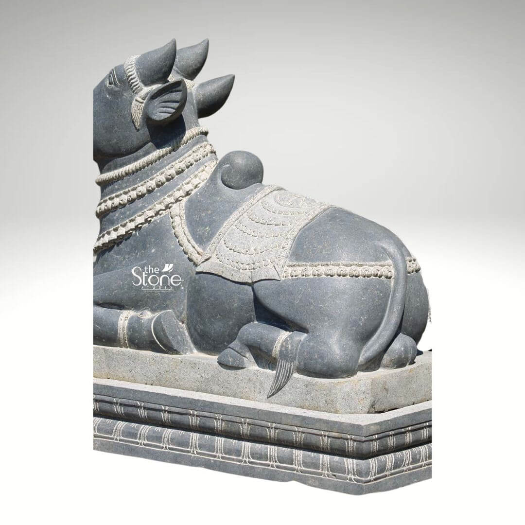 Nandi Statue in Black Designed Stone 2ft 4 A majestic 2-foot Nandi bull statue meticulously carved from black stone is presented from a rear-three-quarter view resting on a multi-tiered pedestal against a plain white background. The statue depicts Nandi in a traditional reclining posture facing away and slightly to the left conveying a sense of calm and steadfast devotion. The black stone has a rich dark gray hue with subtle natural variations and a smooth polished finish that beautifully highlights the intricate details of the sculpture. Nandi is lavishly adorned with elaborate decorative elements all expertly carved into the stone. These include multiple strands of finely beaded necklaces draped around its neck and chest with delicate bells suggested at the bottom. A beautifully patterned saddlecloth covers its back, featuring intricate geometric and traditional designs bordered by a textured band. A ceremonial headpiece is visible between its robust horns and the ears are also finely detailed. A distinct hump is prominent on its back characteristic of the sacred bull. The statue rests on a substantial multi-tiered black stone pedestal which features elegantly carved horizontal lines and traditional ornamental patterns along its edges providing a sturdy and dignified base. The overall impression is one of profound tranquility strength and unwavering devotion making this Nandi bull statue an ideal piece for a home garden, or sacred space reflecting traditional Indian artistry and spiritual significance in its durable black stone form.