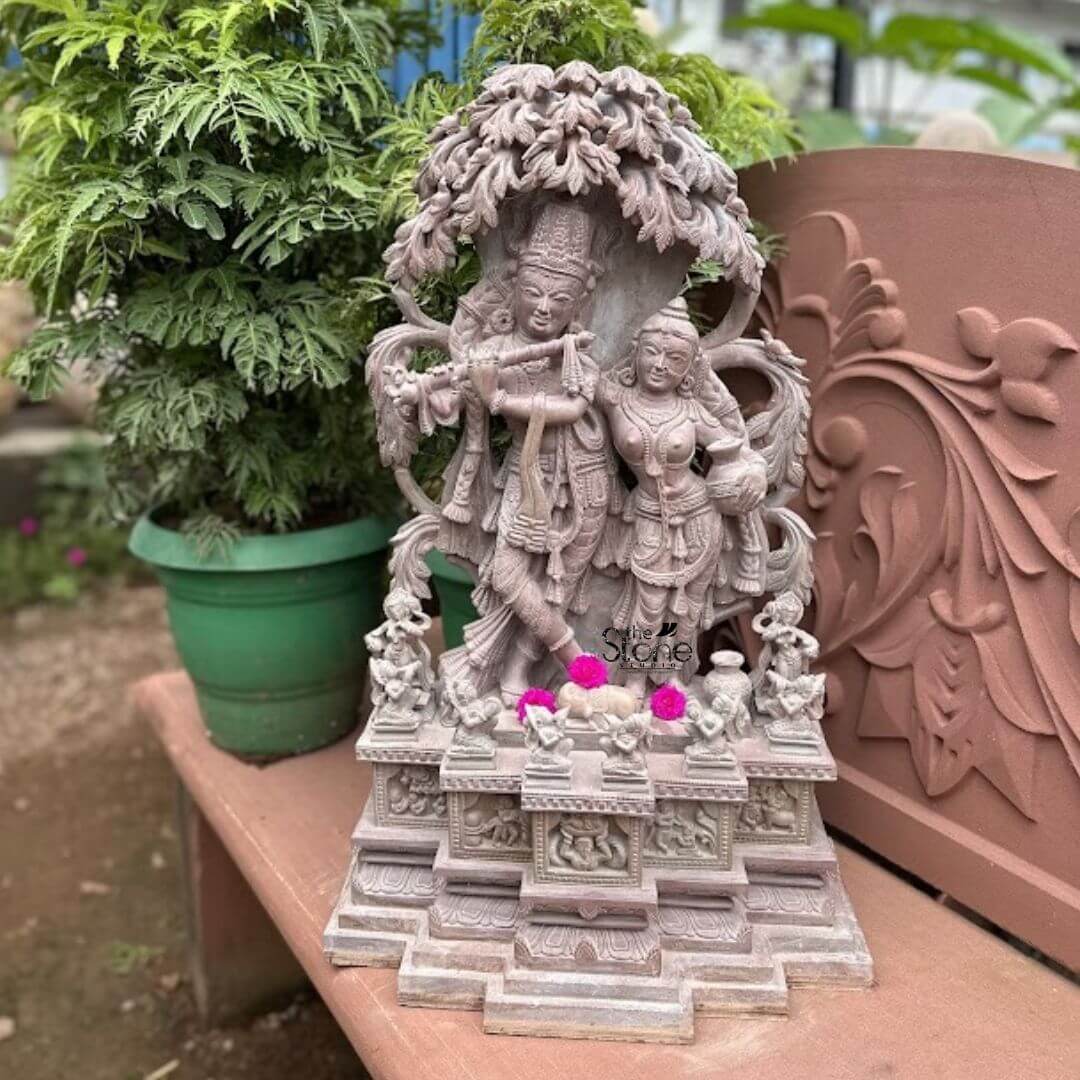 Radha Krishna Statues in Soap Stone 2ft 3 A captivating 2-foot Soapstone Radha Krishna statue perfect as a Radha Krishna Stone Statue for Home is beautifully displayed on a reddish-brown outdoor bench or table amidst a backdrop of lush green plants. The statue carved from light grey soapstone with a subtle reddish tint depicts Lord Krishna and Radha standing lovingly side-by-side under a intricately carved tree. Krishna distinguished by his crown holds a flute to his lips while Radha stands gracefully beside him her hand resting gently on his arm. The craftsmanship is exquisite with intricate details defining their elaborate crowns delicate jewelry and flowing traditional garments. The soapstone's soft texture allows for fine nuanced carving bringing out the tender expressions on their faces and the intricate patterns of their attire. The tree canopy above them is finely sculpted with leaves and branches providing a symbolic shelter and enhancing the romantic setting. The figures stand on a multi-tiered pedestal also carved from soapstone featuring smaller relief sculptures of devotees or mythological scenes adding depth and narrative to the base. Small pink flowers are placed as offerings in front of the deities, indicating a devotional context. The surrounding environment of potted green plants including a large plant in a green pot behind the statue and a carved terracotta panel to the right creates a natural and aesthetically pleasing backdrop. This 2-foot Soapstone Radha Krishna statue is an exquisite example of religious artistry designed to bring blessings love and aesthetic beauty to any home making it an ideal focal point for devotion or decorative accent perfectly aligning with an article focusing on such charming Radha Krishna statues for home interiors.