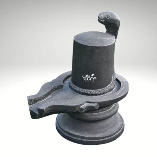 An exquisite 18-inch Shiva Linga sculpture meticulously carved from black granite is presented against a clean softly graded background that transitions from light grey to white. This sacred Hindu icon central to Shaivism is depicted with a highly polished and smooth cylindrical Lingam shaft which symbolizes Lord Shiva's unmanifested energy. Atop the Lingam a finely sculpted five-headed cobra rises its hood spread protectively over the top signifying devotion protection and the cosmic energy associated with Shiva. The texture of the snake's scales is subtly detailed around its base. The Lingam is set within a robust Yoni base which is also crafted from the same dark black granite. The Yoni features a spouted channel on the left side designed to collect and drain offerings poured over the Lingam during worship. The entire structure rests on a circular pedestal providing a stable and aesthetically pleasing foundation. The deep black color and smooth finish of the granite emphasize the sculpture's elegant form and spiritual gravitas making it a striking piece of devotional art. The overall design of this black granite Shiva Linga statue is traditional yet refined perfectly conveying its significance for veneration and its connection to an article discussing such sacred sculptures particularly those made from durable and beautiful black granite.