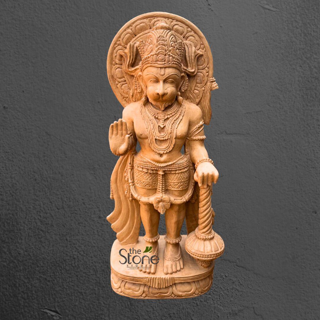 Hanuman Statue in Red Stone Standing Position 3ft 1 magnificently detailed, full-body statue of the Hindu deity Lord Hanuman, expertly carved from a light-colored, possibly sand-colored stone or wood, stands prominently against a plain, dark gray, textured background. Hanuman is depicted in a powerful yet serene standing posture, embodying strength and devotion. His distinctive monkey-like face features a well-defined snout, expressive eyes, and a calm countenance, suggesting wisdom and unwavering loyalty. He wears an elaborately sculpted, multi-tiered crown adorned with intricate traditional patterns. Behind his head, a beautifully carved circular halo (prabhamandala) frames him, signifying his divine aura and spiritual significance. His muscular torso is adorned with multiple layers of ornate necklaces, including a prominent central pendant that rests on his chest. His right hand is raised in the Abhaya Mudra, a gesture of reassurance, protection, and fearlessness, palm facing forward with fingers extended upwards. His left hand rests firmly on the handle of his iconic weapon, a large, spiraled gada (mace), which stands vertically beside him. He is dressed in a richly textured dhoti or lower garment, detailed with folds and decorative patterns, secured by an intricately carved waist belt. His powerful legs are well-proportioned, and his feet rest firmly on a multi-layered, round pedestal. The pedestal itself is a work of art, featuring finely carved lotus petal motifs and other traditional Indian decorative patterns. The material's smooth, polished finish highlights the exceptional craftsmanship and the depth of the carving, showcasing fine details like individual strands in his armlets and the intricate designs on his crown. The statue's light tone stands out vividly against the dark, textured wall behind it, drawing full attention to its intricate details and spiritual presence. A subtle "the Stone Studio" logo is visible at the bottom left of the image, identifying the artisan or source of this devotional sculpture. This exquisite Hanuman idol serves as a powerful representation of courage, loyalty, and strength, and would be a significant spiritual artifact or a central piece in any collection of Hindu religious art or Indian traditional carvings.