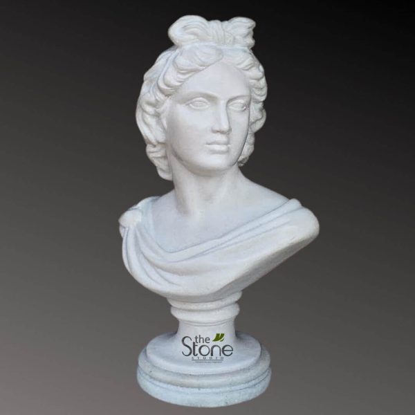 striking, highly detailed white stone bust depicting a classical female figure, likely a goddess or muse from ancient Greco-Roman mythology. The exquisite sculpture captures the head and shoulders of the woman, presented in a three-quarter profile, looking gently towards the viewer's right. Her intricately sculpted hair is styled in soft, flowing waves, gathered elegantly at the back and topped with a prominent, decorative bow or knot, adding a delicate touch to her classical coiffure. She possesses an idealized, serene expression with finely carved features: a straight nose, subtly full lips, and almond-shaped eyes set beneath gently arched brows. Graceful folds of drapery, suggestive of an ancient Greek chiton or Roman toga, are skillfully rendered, cascading over her left shoulder and across her chest, adding a sense of movement and classical elegance to the composition. The pristine white finish suggests a high-quality material such as marble, cast stone, or a similar composite, emphasizing its sculptural form and smooth texture. The bust is supported by a slender, fluted pedestal that gracefully widens into a robust, multi-tiered circular base with elegant moldings. Prominently displayed on the front of the base is the "the Stone STUDIO" logo, featuring elegant script and the tagline "Sculpting Your Imagination," indicating this is a contemporary reproduction or original work produced by an artisan stone studio. The entire sculpture is expertly lit against a muted, gradient dark gray background, which beautifully accentuates its smooth contours and intricate details without distraction. This piece embodies timeless classical aesthetics, offering a perfect representation of ancient artistry and serving as an elegant statement piece for interior design, garden decor, or as a focal point in a collection of historical reproductions, highlighting the craftsmanship available from "the Stone STUDIO."