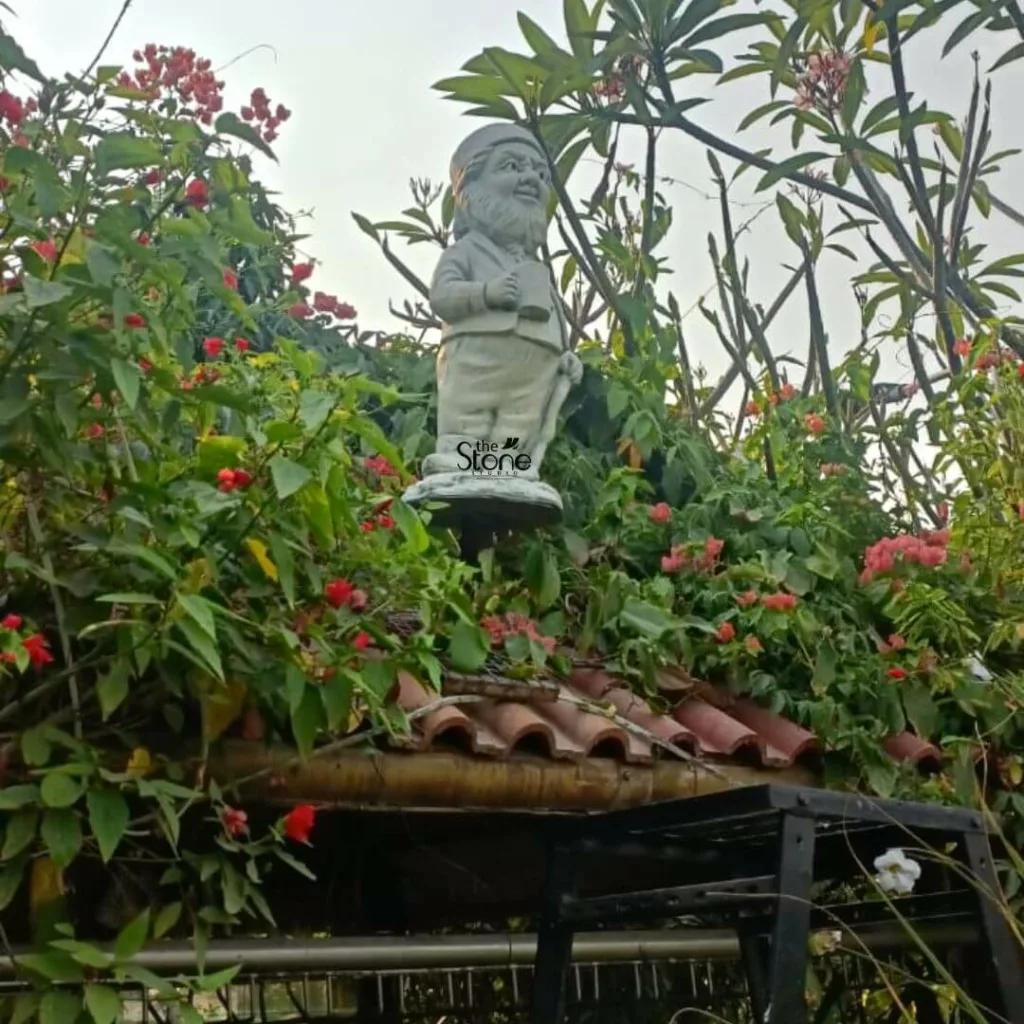 charming, light-colored stone garden gnome statue stands elevated amidst an explosion of lush greenery and vibrant flowers, capturing a serene outdoor garden scene. The gnome, crafted from what appears to be concrete or weathered stone, features a jovial, bearded face with a friendly smile, a conical hat, a jacket, and trousers. He holds a traditional tankard or mug in his right hand and leans gently on a sturdy spade or walking stick with his left, embodying a classic, whimsical garden spirit. The statue is prominently displayed on a circular pedestal, with the brand name "The Stone Studio" subtly integrated as a watermark at its base, hinting at its craftsmanship or origin as a piece of garden art. It is enveloped by a rich tapestry of dense, dark green foliage and abundant clusters of small, fiery red flowers, possibly Ixora, which add a striking pop of color against the verdant background. In the upper right portion of the image, slender branches with sparser green leaves and hints of delicate pale pink flowers, potentially Frangipani (Plumeria), reach towards a soft, bright, and slightly hazy sky. Below the overflowing plant life and the gnome's perch, the textured, reddish-brown curves of traditional terracotta roof tiles are clearly visible, suggesting the statue is placed on a garden shed roof, a decorative gazebo, or an elevated feature within a landscape design. Further down and partially obscured by the foliage, a dark metal structure, possibly a railing or part of a garden stand, can be seen, with a single delicate white flower blooming near it. The overall composition highlights the beauty of natural garden elements combined with ornamental garden decor, showcasing a delightful and rustic aesthetic perfect for enhancing any outdoor space. This detailed image could be used to illustrate an article about garden sculptures, outdoor decorative items, or the artistic offerings of "The Stone Studio."