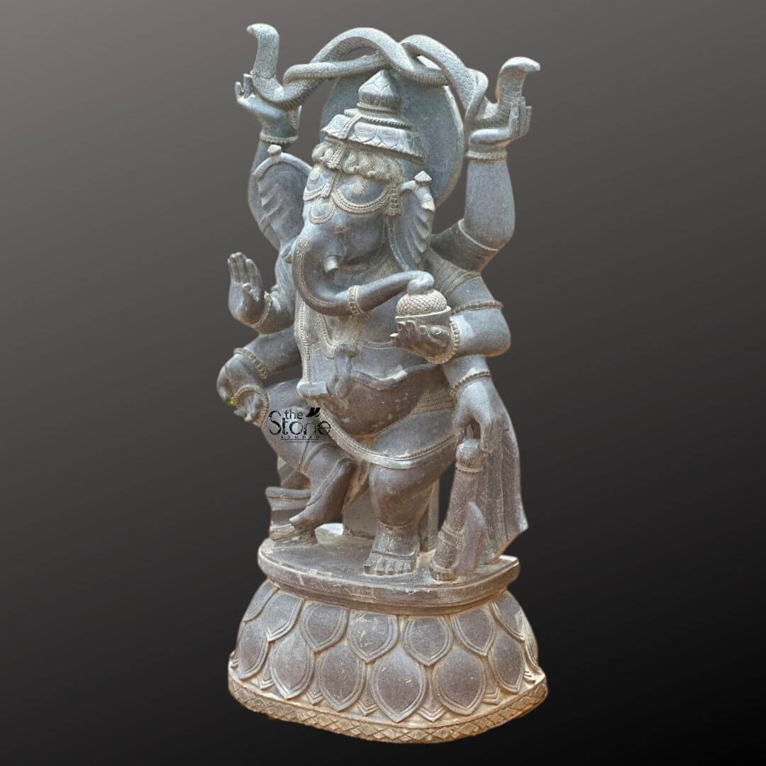 Dancing Ganapati Murti With Snakes 4' 4 A magnificent four-foot-tall hand-carved statue of the Hindu deity Lord Ganesha stands centered against a dark, graduated background showcasing its detailed craftsmanship. Sculpted from a single block of solid black stone the deity is depicted in a dynamic dancing posture known as Nataraja Ganesha. He is captured mid-movement with his left leg gracefully raised and bent across his body while his right foot remains firmly planted on an intricately carved circular lotus pedestal. The statue radiates a sense of joyful rhythm and spiritual grace making it a commanding presence. The four-armed deity is adorned with elaborate traditional ornaments all meticulously carved from the same dark stone. His elephant-head features a serene expression and is crowned with a towering tiered mukuta detailed with decorative patterns. He wears multiple necklaces including a sacred thread armlets wristlets and anklets all with detailed textures. His upper right hand holds a pasha and his upper left hand holds an ankusha. His lower right hand is in the abhaya mudra while his lower left hand holds a bowl of modaks toward which his trunk curls adding a charming life-like detail. The entire sculpture exhibits exceptional artistry with deep relief carvings defining the folds of his flowing dhoti and the intricate patterns on his jewelry and crown. The black stone material has a rich dark matte finish that absorbs light emphasizing the depth and complexity of the carvings. The circular lotus pedestal upon which he dances is also intricately carved with stylized petal layers providing a stable and decorative base for the monumental figure. The statue is presented as a singular object of art highlighting its religious significance and detailed artistry for the article.