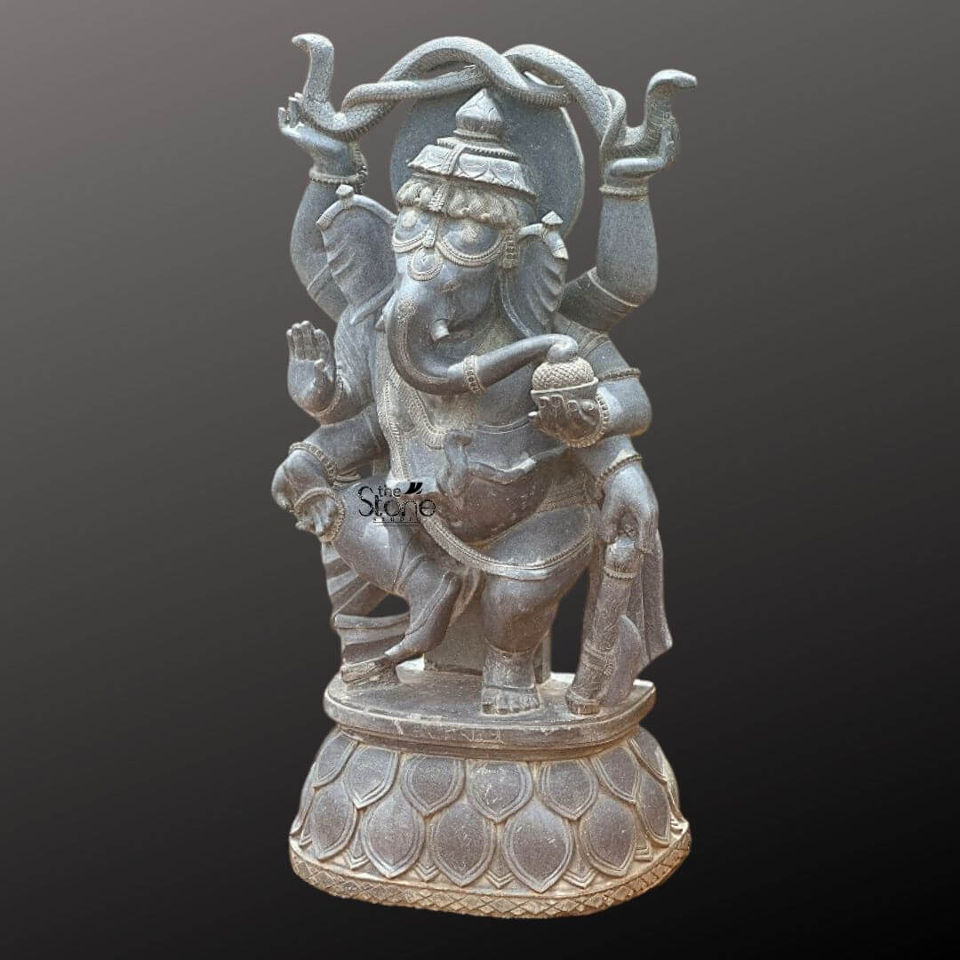 Dancing Ganapati Murti With Snakes 4' 5 A magnificent four-foot-tall hand-carved statue of the Hindu deity Lord Ganesha stands centered against a dark graduated background showcasing its detailed craftsmanship. Sculpted from a single block of solid black stone the deity is depicted in a dynamic dancing posture known as Nataraja Ganesha. He is captured mid-movement with his left leg gracefully raised and bent across his body while his right foot remains firmly planted on an intricately carved circular lotus pedestal. The statue radiates a sense of joyful rhythm and spiritual grace making it a commanding presence suitable for a grand home entryway or temple. The four-armed dancing Ganesha is adorned with elaborate traditional ornaments all meticulously carved from the same dark stone. His elephant-head features a serene expression and is crowned with an ornate tiered mukuta detailed with decorative patterns and a central floral motif. He wears multiple necklaces including a sacred thread armlets wristlets and anklets all with detailed textures that highlight the hand-carved nature of the piece. His upper right hand holds a pasha and his upper left hand holds an ankusha. His lower right hand is in the abhaya mudra while his lower left hand holds a bowl of modaks toward which his trunk curls adding a charming life-like detail to the divine form. The entire sculpture exhibits exceptional artistry with deep relief carvings defining the folds of his flowing dhoti and the intricate patterns on his jewelry and crown. The black stone material has a rich dark matte finish that absorbs light emphasizing the depth and complexity of the carvings and giving the statue a powerful timeless quality. The circular lotus pedestal upon which he dances is also intricately carved with stylized petal layers providing a stable and decorative base for the monumental figure. The statue is presented as a singular object of art highlighting its religious significance and the detailed artistry for the article.