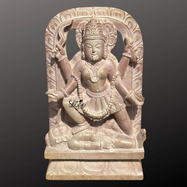 An exquisitely hand-carved 8-inch small Maa Kali statue for home puja, masterfully sculpted from natural pinkstone with a weathered, antique look. The statue captures the Hindu Goddess Kali in her traditional and powerful Dakshina Kali pose, standing with her right foot firmly placed on the chest of a prostrate Lord Shiva, symbolizing the victory of divine energy over ego and the ultimate balance of the universe. The sculpture is rich in Vedic iconography, featuring the Goddess with four arms holding sacred attributes, including a curved sword (khadga) and a severed head, representing the destruction of ignorance. She is meticulously detailed with a protruding tongue, a towering tiered crown (mukut), layered necklaces, and a skirt made of human arms. Framing the central figure is a grand, circular Prabhavali (ornate arch) embellished with intricate traditional floral and geometric relief patterns. The natural pinkstone material exhibits a rustic, grainy texture with earthy shades of rose and tan, giving the piece a timeless, heritage feel that perfectly complements a traditional home mandir. Resting on a sturdy tiered pedestal with lotus-petal motifs, this compact 8-inch stone Maa Kali idol is a perfect spiritual centerpiece for a personal altar or a small meditation corner, designed to invite protection, strength, and transformative energy into any sacred living space.