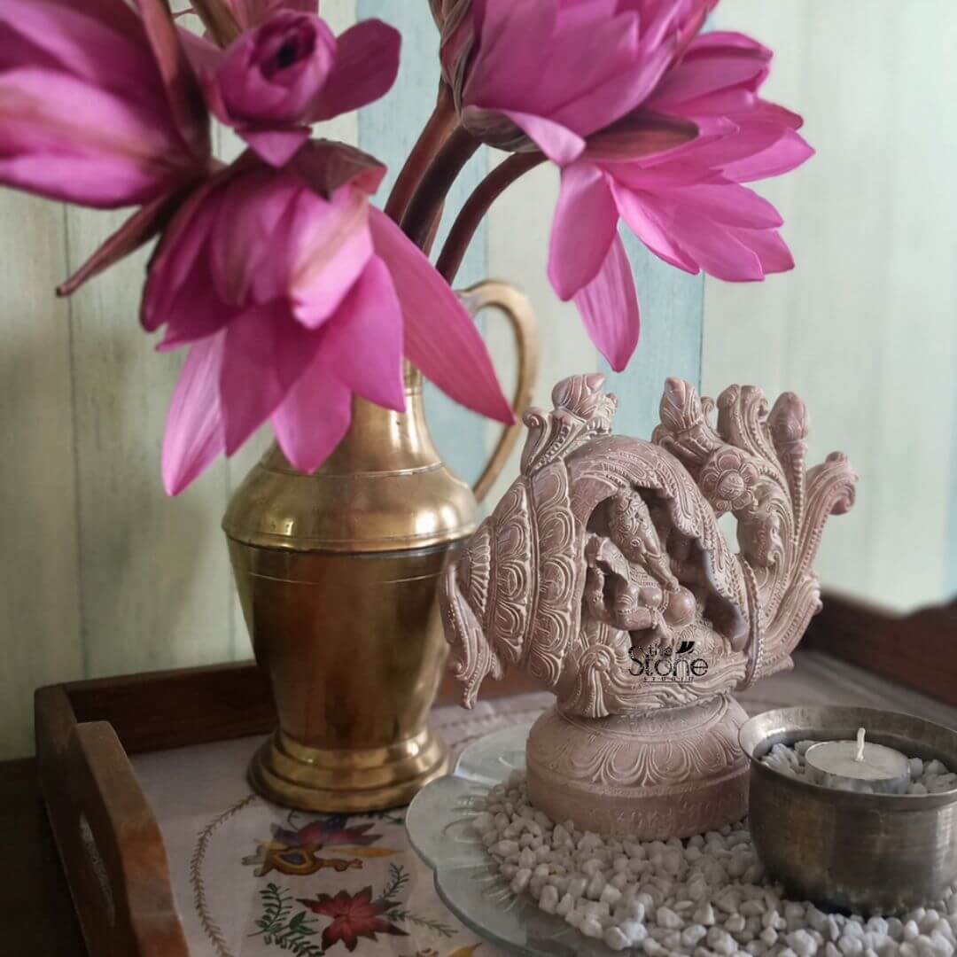 Ganpati Idol Gift 8" 2 A beautifully crafted eight-inch-tall statue of Lord Ganesha hand-carved from a warm pink-hued soapstone is the central focus of this decorative indoor arrangement. The Ganesha statue is uniquely integrated within a stylized shankh form symbolizing a blend of auspicious elements. The deity's serene elephant head and four-armed body are meticulously carved in deep relief nestled within the conch's opening. The conch shell itself is intricately detailed with traditional Indian ornamental patterns and the entire piece rests upon a matching carved circular pedestal showcasing the artisan's skill in rendering fine detail from the soft soapstone. The soapstone sculpture sits on a small glass plate covered in tiny white river stones adding texture and a clean base to the display. To the left of the statue stands a tall vintage brass kalash or water pot its polished surface showing a warm patina and reflecting the ambient light. This brass pot holds a vibrant lush arrangement of several large deep-pink lotus flowers on long stems providing a striking color contrast to the pink stone and brass. The entire ensemble is presented on a dark wooden serving tray which rests on a textured surface possibly a woven table runner with subtle embroidered floral patterns visible beneath the objects. The background is softly blurred featuring a light neutral-toned wall with a faint greenish tint creating a calm and uncluttered backdrop that emphasizes the objects on the tray. The lighting is soft natural daylight filtering from the side casting gentle shadows that highlight the rich textures of the hand-carved soapstone the hammered brass and the delicate petals of the lotus flowers. This composition creates a harmonious and spiritually uplifting vignette perfectly illustrating the statue's suitability as a sophisticated and meaningful piece for home decor. A small lit diya in a hammered brass cup is placed to the right of the Ganesha conch adding a warm inviting glow to the scene.