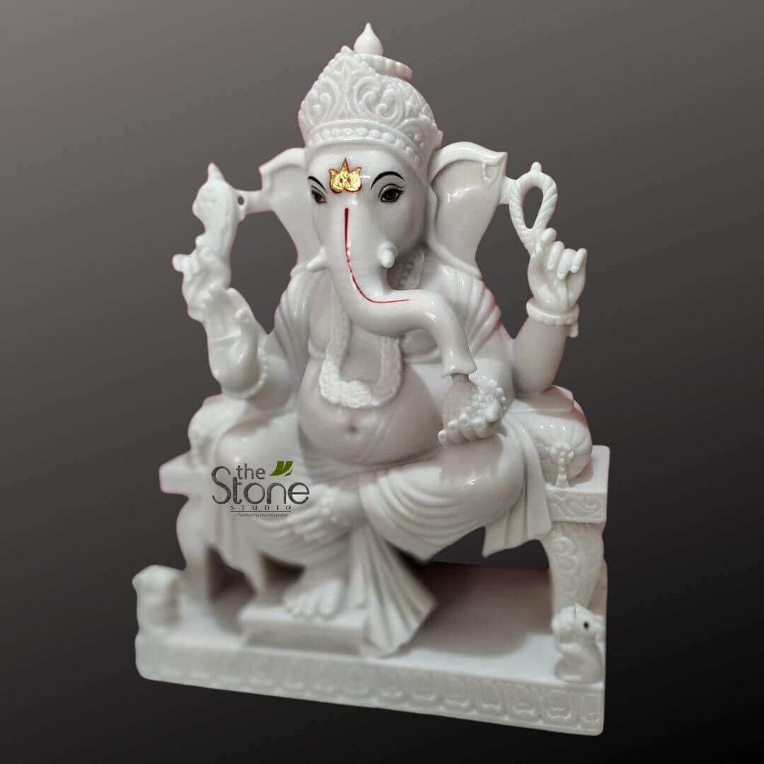 Ganesha Marble Statue 30" 7 stunning, pristine white marble sculpture of Lord Ganesha, the elephant-headed Hindu deity, is showcased against a plain, dark grey gradient background that emphasizes its radiant polish and intricate details. The statue depicts Ganesha seated gracefully in Lalitasana, a relaxed posture with one leg folded and the other gently resting on the ground, on an ornately carved throne or asana. Ganesha's majestic elephant head faces slightly towards the viewer, featuring large, expressive dark eyes, and a trunk that curves gently to his left (the viewer's right). A distinctive red tilak mark is painted along the curve of his trunk, and a golden, stylized leaf or flower emblem adorns his forehead, adding a touch of regal elegance. He wears an elaborately carved crown with swirling patterns, signifying his divine status. The deity is depicted with four arms, holding traditional attributes. His upper right hand grasps an ankusha (elephant goad), while his upper left hand holds a paasha (noose). His lower right hand rests near his lap, delicately holding a modak (sweet dumpling), symbolizing blessings and wisdom. His lower left arm is bent, with the hand in a gesture of repose, possibly holding his broken tusk, representing the sacrifice he made while writing the Mahabharata. Ganesha's characteristic pot-belly is prominent, symbolizing abundance and prosperity, and a simple, delicate garland is draped around his neck. The drapery of his dhoti is subtly carved, suggesting soft folds and movement. The ornate throne on which Ganesha sits features decorative carvings on its legs and backrest, enhancing the overall grandeur. At the base of the throne, near his right foot (viewer's left), his vahana (mount), a small, intricately carved mouse (Mushika), is visible, looking up at him devotedly. The rectangular base of the entire statue also features continuous, delicate relief carvings, adding to the craftsmanship. This exquisite Ganesha idol, likely a masterpiece from a stone carving studio, radiates a serene aura, making it ideal for spiritual decor, a home altar, or as a powerful symbol of wisdom, success, and the removal of obstacles. The smooth, glossy finish of the white marble highlights the meticulous hand-carved artistry, presenting a timeless piece of Indian religious art. The logo "the Stone STUDIO" is subtly visible at the bottom left, indicating the source of this fine marble sculpture.
