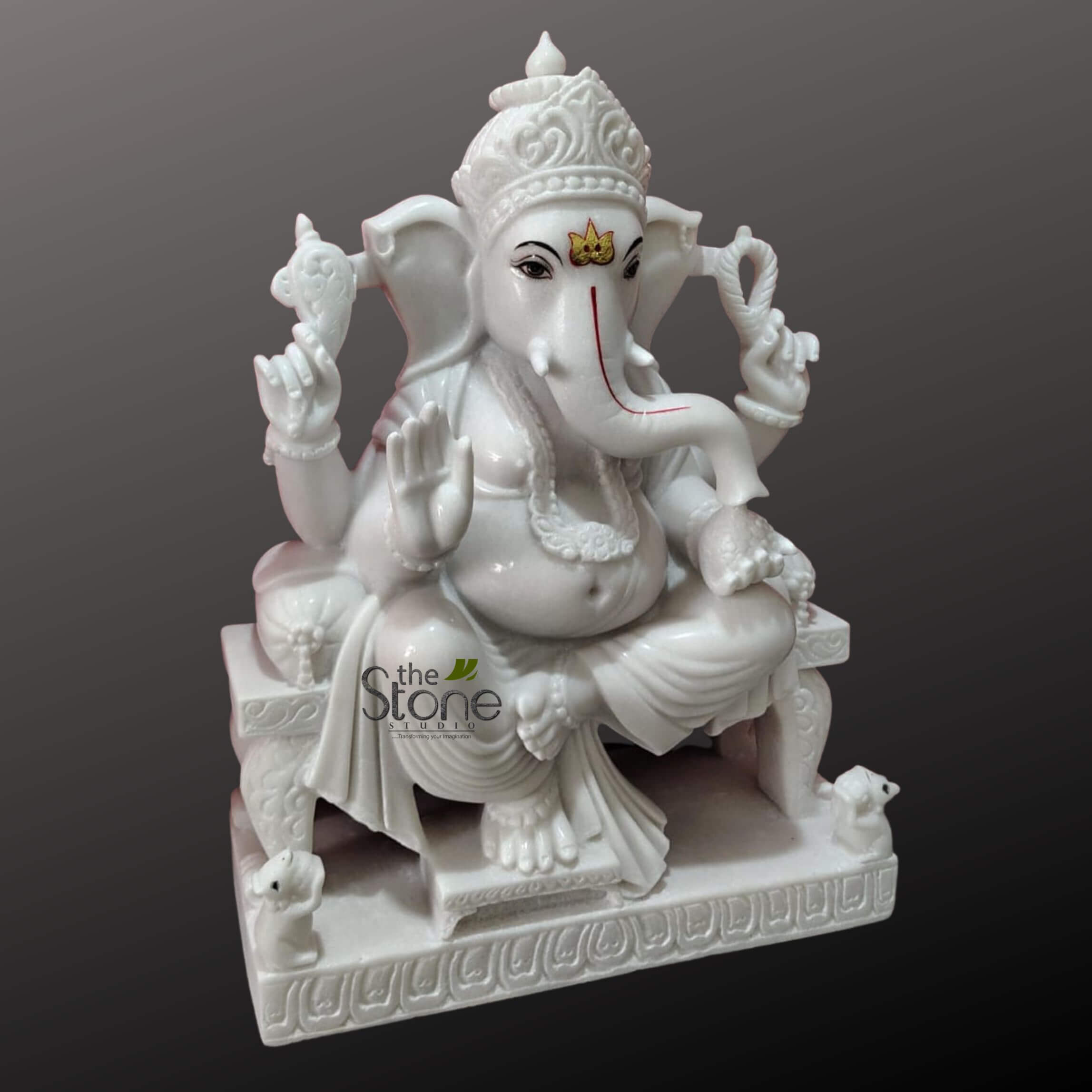 Ganesha Marble Statue 30" 6 stunning, intricately carved white marble statue of Lord Ganesha is prominently displayed against a plain, dark gray gradient background. The divine elephant-headed deity is seated in a regal, relaxed posture, with one leg folded and the other slightly raised, on a highly ornate, multi-tiered pedestal of matching white marble. Ganesha's benevolent face features large, fan-like ears, expressive black eyes, and a golden, crown-like tilak adorning his forehead. His prominent trunk is gracefully curled to his left, marked with a distinctive red tilak line. One tusk is visibly broken, an iconic characteristic. He wears an elaborate, multi-tiered crown with delicate carvings and various sculpted adornments including a detailed necklace, armlets, and anklets. His portly belly, symbolizing prosperity and abundance, is covered by flowing robes that show intricate folds and drapes, adding to the statue's dynamic feel. Each of his four arms holds a symbolic attribute: his upper right hand firmly grasps an ankusha (goad), while his upper left hand holds a pasha (noose). His lower right hand is raised in the Abhaya Mudra, a gesture of blessing and fearlessness, offering protection to devotees. His lower left hand gently holds a sweet modak (ladoo), his favorite treat. The sturdy, rectangular pedestal beneath Ganesha is also crafted from white marble, featuring finely sculpted, continuous relief carvings of what appear to be floral or geometric patterns along its front and side edges. Small, intricately carved figures of Mushak, Ganesha's divine vehicle (a mouse), are positioned at the front corners of the base, looking up towards the deity. The pristine white, polished marble of the entire sculpture brilliantly stands out against the dark, neutral backdrop, highlighting the exquisite craftsmanship and devotion captured in this traditional Hindu deity art piece, suggesting it could be a centerpiece for home decor, spiritual practices, or an article about sacred sculptures and artisanal stone work.