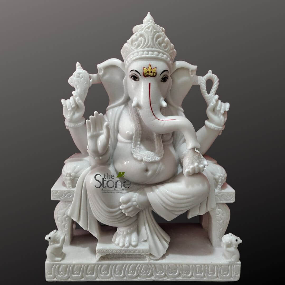 Ganesha Marble Statue 30" 5 stunning, exquisitely carved white marble sculpture of Lord Ganesha, the elephant-headed Hindu deity, is presented centrally against a plain, dark gray gradient background that enhances its pristine appearance. The statue is meticulously crafted with a smooth, polished finish, highlighting the intricate details of the stone. Lord Ganesha is depicted in a serene, regal seated posture, likely Lalitasana (one leg drawn up, the other folded), on an ornately sculpted pedestal or throne. His iconic elephant head features large, fanned ears and expressive, dark eyes. A traditional golden crown motif (mukuta) with a red tilak adorns his forehead, while his trunk curls gracefully to his left, outlined with a delicate red line. He wears an elaborate, multi-tiered crown (mukut) intricately carved with traditional designs, symbolizing his divine royalty. The deity has a prominent, rounded pot belly, signifying prosperity and abundance, and is adorned with numerous carved ornaments including necklaces, armlets, bracelets, and anklets, all rendered in the same luminous white marble. Flowing drapery with delicate folds cascades from his waist, adding to the sculpture's realistic texture and elegance. Ganesha is depicted with four arms, a common representation of his omnipotence. In his upper right hand, he holds an ankusha (goad), symbolizing spiritual awakening and control. His upper left hand holds a pasha (noose), representing the binding nature of attachments. His lower right hand is raised in Abhaya Mudra, offering blessings, protection, and reassurance to devotees. In his lower left hand, he holds a small, round sweet delicacy, traditionally a modak or laddu, symbolizing the rewards of spiritual pursuit and inner joy. The intricately carved base features a decorative border of continuous wave or floral patterns. Flanking the front of the pedestal are two small, exquisitely detailed mooshaks (mice), Ganesha's traditional vehicle, looking up towards the deity. This magnificent Hindu religious idol exemplifies exceptional traditional Indian stone carving artistry, making it an ideal piece for spiritual contemplation, home altars (puja rooms), or as a significant decorative element embodying wisdom, prosperity, the remover of obstacles, and auspicious beginnings. The presence of "the Stone Studio" watermark suggests this is a high-quality, handcrafted spiritual art piece.