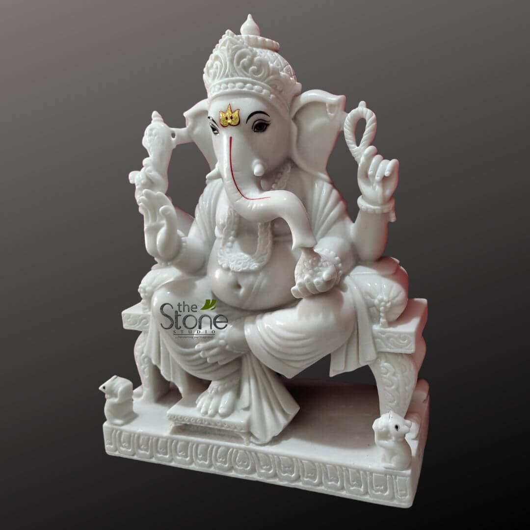 Ganesha Marble Statue 30" 4 majestic and highly detailed white marble statue of Lord Ganesha, depicted in a seated, relaxed pose (often called Lalitasana or a similar posture), against a plain, soft gray background. The pristine white stone, with its smooth, polished finish, highlights the intricate carvings. Ganesha, the elephant-headed Hindu deity of wisdom, prosperity, and good fortune, is shown with his characteristic large ears, a prominent pot belly, and four arms. His trunk curls gracefully to his left, touching a modak (sweet dumpling) held in his lower left hand. A vibrant red tilak (sacred mark) extends down his trunk, and a golden sacred symbol, possibly an 'Om' or a specific tilak, adorns his forehead. His large, serene eyes gaze forward. He wears an ornate, multi-tiered crown (mukut) on his head, and intricately carved jewelry adorns his body, including necklaces, armlets, wristlets, and a sacred thread (yajnopavita) draped diagonally across his chest. His garment, a dhoti, is elegantly draped over his legs, showing finely sculpted folds. In his upper right hand, Ganesha holds a paasha (noose or rope), symbolizing the attachment to worldly desires, while his upper left hand grasps an ankusha (goad), representing the control of desires. His lower right hand is either raised in a gesture of blessing (Abhaya Mudra) or gently holds another modak, completing the traditional attributes. The deity sits upon an intricately carved throne-like pedestal. The base of the pedestal features a continuous pattern of decorative motifs, possibly lotus petals or a geometric design, indicating exceptional craftsmanship. On either side of the base, two small, attentive mooshak (mice), Ganesha's traditional vehicle (vahana), are carved looking up towards the deity, adding to the spiritual significance of the piece. A subtle watermark of "the Stone STUDIO" is visible in the lower-left area of the statue, indicating its origin or artisan. This exquisite marble idol embodies divine grace and devotion, perfect for a spiritual decor collection, home temple, or as a focal point for Hindu worship and meditation, reflecting the rich heritage of Indian religious art and sculpture.