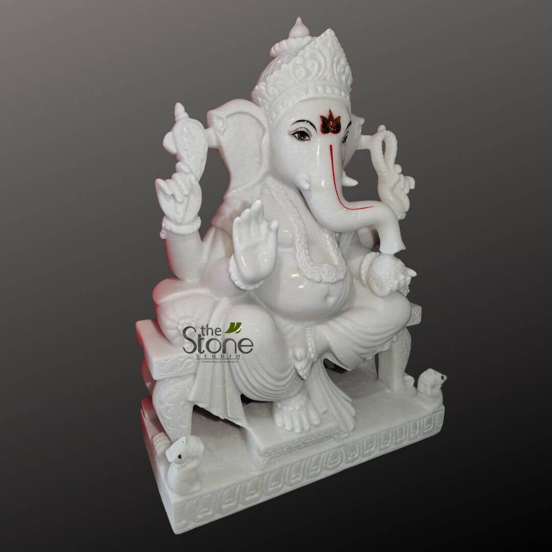 Ganesha Marble Statue 30" 2 magnificent, intricately carved white marble statue of Lord Ganesha, seated gracefully in a relaxed pose (Lalitasana) on an ornate, multi-tiered pedestal. The idol is crafted from pristine white marble, giving it a luminous and pure appearance, with subtle red-orange accents defining his features. Ganesha's elephant head features prominent, fanned ears and serene dark eyes, outlined in black, with a distinctive red tilak mark adorning his forehead. His trunk curves gently to the left, showcasing a vibrant red line that runs along its length, indicating an auspicious mark. He wears an elaborately carved, multi-tiered crown and is adorned with intricate marble jewelry, including a necklace, armlets, wristlets, and a sacred thread (Yajnopavita) draped diagonally across his pot-bellied torso, symbolizing prosperity and spiritual dedication. The deity is depicted with four arms. His upper right hand holds a pasha (noose), while his upper left grasps an ankusha (elephant goad). His lower left hand delicately holds a modak (sweet dumpling), symbolizing wisdom and rewards, and his lower right hand is raised in the traditional Abhaya Mudra, a gesture of fearlessness and blessing. His lower body is covered by finely detailed, flowing drapery with intricate pleats and folds. Around the elaborately carved base of the statue, several small, endearing figures of Mushika, Ganesha's mouse vahana (mount), are visible, some looking up at their lord. The entire sculpture is set against a smooth, graduated gray background that accentuates the purity and detailed craftsmanship of the white marble. The logo "the Stone Studio" is visible in the lower left, indicating the source of this exquisite piece of art. This highly detailed and auspicious Ganesha idol embodies spiritual devotion, skilled craftsmanship, and serves as a perfect representation for discussions on Hindu religious decor, marble sculptures, home temple idols, or Indian traditional art.
