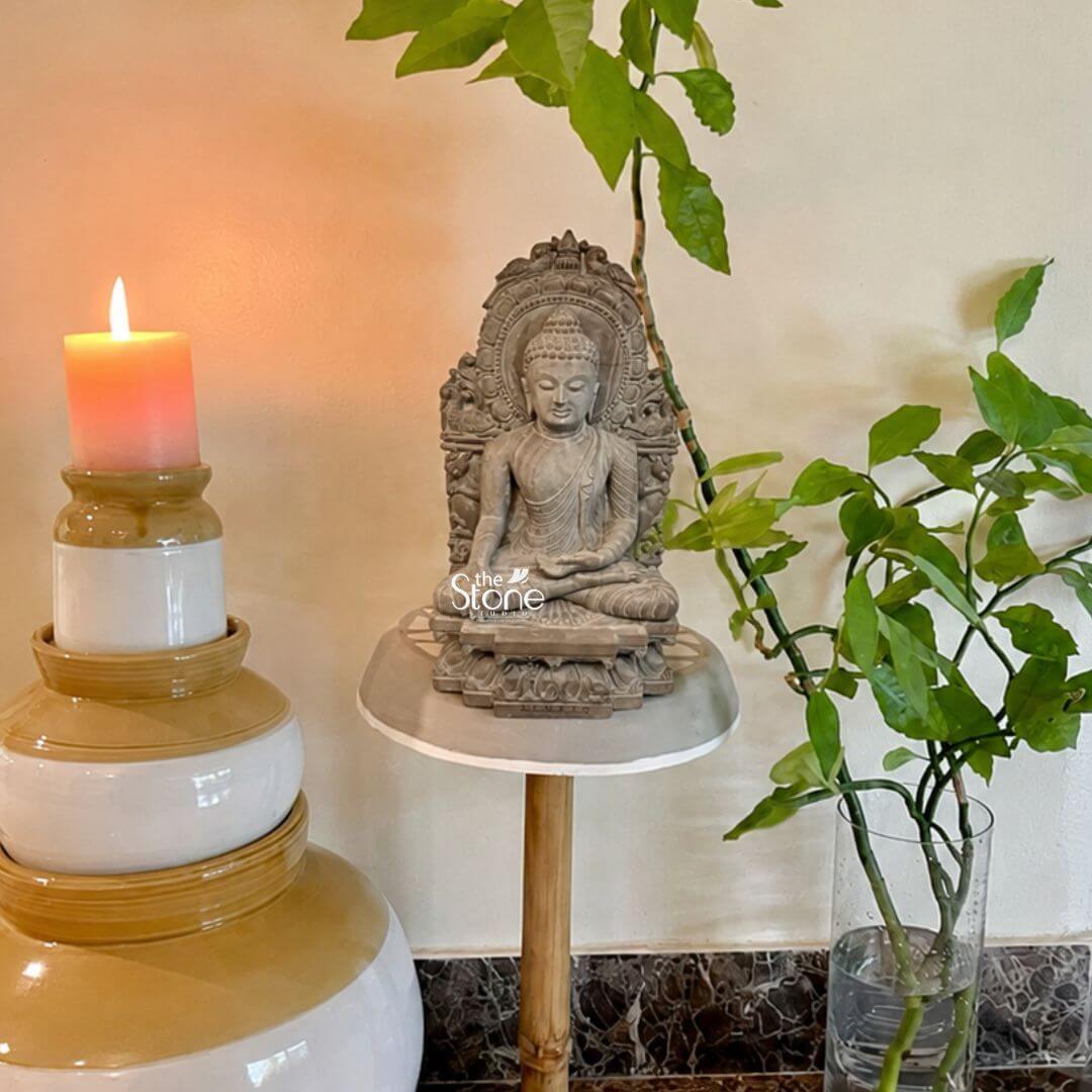 Buddha Statue 8" 3 An exquisite 8-inch Buddha idol carved from pink stone serves as a focal point in a beautifully arranged home decor setting. The Buddha is depicted in a traditional meditative seated posture with his hands gently resting in his lap emanating a profound sense of peace and contemplation. His expression is serene with downcast eyes symbolizing inner reflection and wisdom. The statue is placed on a round light-colored platform supported by a slender wooden stand. The pink stone Buddha statue features intricate carvings particularly on the elaborate aureole or throne back that frames the deity. This backdrop is adorned with detailed motifs possibly depicting foliage or celestial beings adding to the idol's sacred aesthetic. The Buddha's hair is meticulously sculpted in tight curls and his robes are rendered with delicate folds. The warm subtle pink hue of the stone adds a gentle and inviting quality to the piece. To the left of the statue a tall tiered candle with a lit flame casts a soft inviting glow creating a tranquil ambiance. This 8-inch pink stone Buddha idol is an ideal subject for an article on small Buddha statues for home decor. To the right a vase with lush green plant cuttings adds a touch of natural freshness complementing the serene theme. The thoughtful arrangement of the statue the candle and the plants against a neutral-toned wall with a darker tiled dado creates a harmonious and calming display perfect for enhancing the spiritual and aesthetic appeal of any home interior.