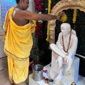 captivating scene unfolds as a devotee or priest performs a sacred Hindu ritual (Abhishekam) on a meticulously crafted white marble statue of Shirdi Sai Baba. The man, with dark skin and a partially visible shaved head and shoulder, is dressed in a vibrant yellow dhoti, traditional South Indian attire, featuring a subtle black, white, and red striped border. He stands reverently on the left, holding a light green coconut with both hands, carefully tilting it to pour a liquid offering – likely water or milk – over the head of the seated deity. The exquisite white marble statue of Sai Baba sits centrally on a simple, matching stone pedestal. Sai Baba is depicted in his iconic meditative pose, with one leg drawn up and the other slightly lower, hands resting gently in his lap. He wears a serene expression, a long strand of brown rudraksha beads around his neck, and a distinct red 'tilak' mark on his forehead, signifying spiritual auspiciousness. Behind the statue, the backdrop is a lush green wall of artificial foliage or creeper, adorned with vibrant clusters of red and yellow flowers, creating a natural and sacred ambiance. Framing the deity is an ornate, intricately carved golden archway, also known as a 'prabhavali,' which accentuates the statue's divine presence. Yellow marigold garlands are draped gracefully over both the arch and the green foliage. On the white platform at the base of the statue, several ritualistic items are visible: a silver-colored bucket filled with water, two smaller copper-toned metal cups, and scattered red flower petals, all indicating an ongoing worship ceremony (puja). The entire setting evokes a strong sense of devotion, peace, and spiritual tradition. In the bottom right corner, a subtle watermark reads "The Stone Studio," suggesting the image highlights the fine craftsmanship of the marble idol, connecting to themes of custom stone deity manufacturing, temple artistry, or religious iconography for home shrines and temples. This detailed portrayal emphasizes the significance of high-quality marble Sai Baba idols in contemporary Hindu spiritual practices.