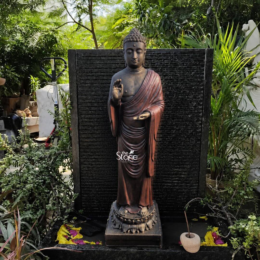 Standing Buddha 5' 3 A medium shot captures a majestic standing Buddha statue expertly crafted from durable weather-resistant fibre positioned as a serene focal point within a lush outdoor garden. The statue is depicted in a traditional standing posture known as Abhaya Mudra with its right hand raised in a gesture of fearlessness and blessing while its left hand is held out palm upwards. The figure's face conveys a profound sense of inner peace and compassion with eyes half-closed and a gentle smile framed by the characteristic snail-shell curls and the ushnisha protrusion on its head symbolizing wisdom and enlightenment. The Buddha figure is finished in a rich dark bronze-like patina on the exposed head and body contrasting elegantly with the flowing reddish-brown hue of its robe or sanghati which hangs in graceful natural folds that accentuate the statue's tall form. The fibre material exhibits a finely detailed texture that mimics traditional metal or stone casting giving the large idol a solid and substantial presence. The statue stands centered upon a decorative multi-layered lotus pedestal providing a stable and sacred foundation. The setting is a tranquil garden alcove with the Buddha statue placed directly in front of a dark textured black stone water wall feature which has subtle horizontal striations. The sound of water cascading down the wall adds to the peaceful atmosphere. The statue is surrounded by a dense variety of thriving green plants and foliage creating a natural enclosure that isolates the sacred space. On the dark stone ledge at the statue's base a few colorful fallen yellow and purple flower petals are scattered along with a smooth rounded stone adding a natural and organic touch to the altar area. Natural daylight filters through the leaves above casting soft shadows that emphasize the statue's contours and rich colors making it a captivating element for garden decor.