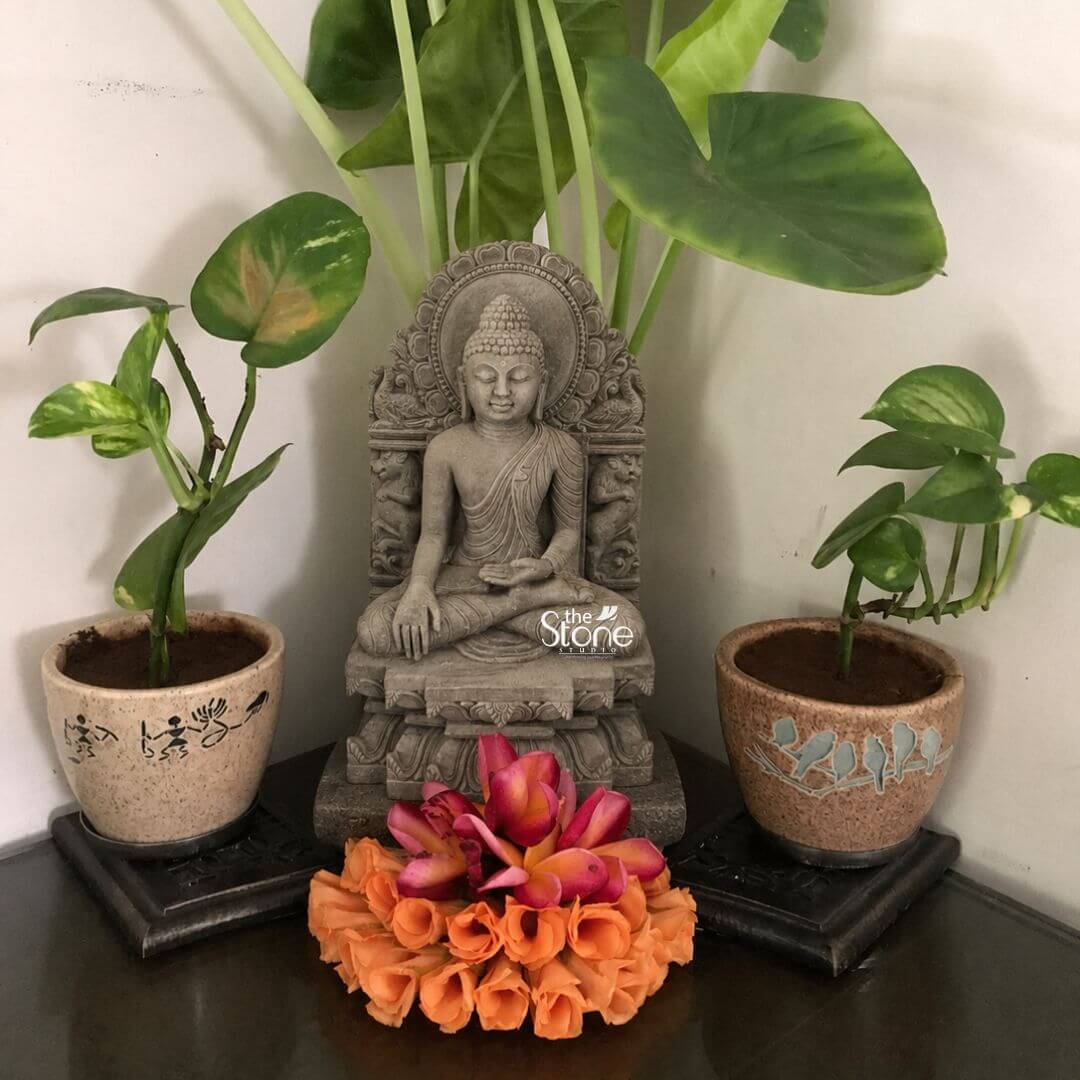 Buddha Sculpture 8" 3 An exquisite 8-inch Small Buddha Home Decor Stone Statue carved from pink stone is beautifully arranged as a serene centerpiece on a dark corner shelf flanked by vibrant potted plants. The Buddha is depicted in a traditional meditative seated posture with his hands gently resting in his lap likely in Dhyana Mudra symbolizing profound concentration and spiritual peace. His expression is serene with downcast eyes a subtle smile and elongated earlobes conveying deep wisdom and tranquility. The pink stone Buddha statue features intricate carvings particularly on the elaborate aureole or throne back that frames the deity. This backdrop is adorned with detailed motifs adding to the idol's sacred aesthetic and artistic richness. The Buddha's hair is meticulously sculpted in tight curls culminating in the ushnisha and his robes are rendered with delicate pleats and folds. The subtle warm pink hue of the stone adds a gentle and inviting quality to the piece. In front of the statue a vibrant floral offering of orange and pink blossoms is artfully arranged adding a pop of color and a devotional touch. This 8-inch pink stone Buddha statue is a perfect subject for an article on small Buddha home decor stone statues. It is flanked by two potted plants with green foliage adding a natural and calming element to the display. The rustic terracotta pots with decorative patterns on either side of the statue further enhance the aesthetic. The entire arrangement creates a harmonious and peaceful focal point ideal for enhancing the spiritual and aesthetic appeal of any home interior fostering a sense of calm and mindfulness.