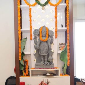 A majestic 3-foot gray stone statue of Lord Vishnu stands as the central deity in a beautifully adorned home altar, radiating a sense of devotion and spiritual presence. The statue depicts Vishnu in a standing posture, holding his traditional attributes in his four hands: the Shankha (conch) and Chakra (discus) in his upper hands, and the Gada (mace) and Padma (lotus) in his lower hands, though the lower right hand's object is less clear. He is adorned with a traditional crown, intricate jewelry, and a flowing dhoti, all carved with meticulous detail from the stone. A vibrant orange marigold garland is draped around his neck, further enhancing his divine appearance. The altar itself is a modern, dark wood and white shelving unit, elegantly decorated for worship. The shelves are framed by vertical strings of bright orange marigold garlands, with a green garland draped across the top, adding a festive and sacred ambiance. Behind the statue, a luminous white panel features intricate lotus-cut designs, which are backlit, creating a soft, reverent glow. Large green banana leaves flank the statue on either side, symbolizing auspiciousness. On the lower shelf in front of Vishnu, a dark tray holds offerings. Various other smaller deities and religious items are visible on the surrounding shelves, completing the sacred space. The overall image captures a beautifully maintained and dedicated prayer area, ideal for a 3-foot Vishnu statue within a home.