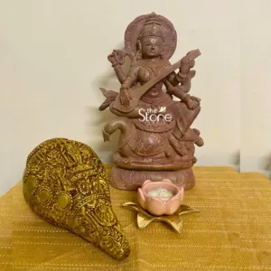beautifully crafted, reddish-brown sandstone statue of Goddess Saraswati, the Hindu deity of knowledge, music, art, speech, wisdom, and learning, is the central and most prominent feature of this image. She is depicted seated serenely and gracefully playing the veena, her iconic stringed instrument, held by her two main arms. The statue showcases her traditional four arms; in addition to holding the veena, one upper right hand holds a mala (rosary), and one upper left hand holds a pustaka (book or scriptures), symbolizing spiritual knowledge and wisdom. Saraswati is seated on her vahana (vehicle), a majestic swan (hamsa), which rests upon a multi-tiered lotus pedestal, all intricately carved from the same reddish stone. Detailed carvings highlight her elaborate crown, jewelry, flowing attire, and the delicate feathers of the swan, reflecting exquisite Indian craftsmanship and traditional artistry often found in spiritual sculptures. To the left of the Saraswati idol lies an equally ornate, elongated object, crafted from what appears to be gold-painted brass or another polished metal. Its surface is richly embossed and engraved with traditional Indian motifs, including small figures of deities or mythological scenes, intricate floral patterns, and circular medallions, giving it the appearance of a highly stylized conch shell (shankha) or a ceremonial vessel. This piece exemplifies detailed metalwork and traditional Indian decorative art. On the right side, a delicate pink ceramic lotus-shaped candle holder cradles a lit white tea light candle, resting on a golden metallic base shaped like lotus leaves. The warm, flickering flame adds a soft, devotional glow to the tableau. All these spiritual and decorative items are arranged thoughtfully on a textured, mustard-yellow or golden-hued woven fabric, possibly a traditional table runner made of jute or a similar natural fiber, which provides a rich backdrop. The background consists of a plain, light beige wall. A "the Stone Studio" watermark subtly overlays the central statue, indicating the origin or specialized material of the stone sculpture. The overall composition evokes an ambiance of peace, devotion, and artistic reverence, highlighting traditional Indian cultural elements and spiritual home decor, suitable for articles discussing Hindu deities, stone carvings, Indian handicrafts, or devotional art pieces.