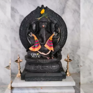 highly detailed and serene photograph showcases a magnificent, meticulously hand-carved black stone sculpture of the Hindu deity Lord Ganesha. The robust idol depicts Ganesha seated in a graceful, relaxed posture, possibly lalitasana, on a multi-layered lotus pedestal, all carved from the same dark, polished material, which appears to be black granite or basalt. His iconic elephant head features large, prominent ears, and his trunk is gently curled to the left, adorned with a reddish-orange tilak mark on his forehead. The deity's four arms are visible; his lower right hand is raised in abhaya mudra, a gesture of fearlessness and blessing, while his lower left hand gently rests, possibly holding a modak (sweet). His upper hands hold traditional attributes like an ankusha (goad) and a pasha (noose), though the intricate details are subtly rendered against the dark stone. A grand, intricately carved prabhavali (decorative arch or backrest) forms a striking halo behind the deity, framing him with traditional Indian mythological or floral motifs, showcasing exceptional artisan craftsmanship in stone carving. The Ganesha idol is adorned with fresh puja decorations: a vibrant yellow marigold flower and green foliage rest delicately on his crown, and a thin white yagnopavitam (sacred thread) drapes across his pot-belly chest. Two bright, triangular pieces of fabric, strikingly striped in hues of pink, green, blue, and yellow with shimmering gold scalloped borders, are draped gracefully over his shoulders, adding a vivid splash of color and festive appeal to the dark, monochromatic sculpture. The entire Ganesha sculpture rests firmly on a clean, multi-tiered white marble platform or pedestal, providing a luminous base. Symmetrically placed on either side of the statue are two elegant, tall brass oil lamps (diyas) with slender, ornate stands, traditionally used in Hindu worship and aarti ceremonies. The background is a simple yet elegant white marble wall with subtle grey veining, which provides a serene and contrasting backdrop, making the dark, polished statue stand out prominently. This image powerfully conveys traditional Indian religious art, devotion, and the exquisite skill of artisanal stone carving, making it ideal for articles on Hindu festivals like Ganesh Chaturthi, spiritual home decor, traditional craftsmanship, or specialized sculpture studios like 'the Stone Studio' (as indicated by the watermark).