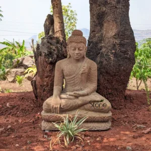 serene and exquisitely carved sandstone Buddha statue is prominently featured in a vibrant outdoor garden setting, embodying peace and tranquility. The handcrafted sculpture depicts Buddha seated in a traditional meditative lotus posture (Padmasana), with both hands resting gently in his lap in the Dhyana mudra, symbolizing deep concentration and spiritual enlightenment. Crafted from natural, earthy-toned stone, the statue has a subtle, granular texture that highlights the intricate details of his softly draped monastic robes, elongated earlobes, gently closed, peaceful eyes, and a subtle, serene smile. His hair is styled in tightly coiled curls, topped with an ushnisha, the cranial bump of spiritual wisdom. The Buddha statue sits firmly on a beautifully detailed multi-tiered lotus pedestal, positioned directly in front of the massive, dark, deeply textured trunks of two ancient trees, creating a harmonious blend with nature. The immediate ground beneath the statue is composed of reddish-brown, dry earth, from which a small, variegated green and cream plant thrives directly in front of the statue's base, adding a touch of verdant life. The background reveals a lush, green landscape, featuring various tropical plants, hints of distant rolling hills or mountains under a bright, clear sky, emphasizing the natural and calming environment. This outdoor garden sculpture serves as a compelling focal point, designed to invite contemplation and foster a sense of inner peace. A subtle "the Stone" watermark, accompanied by a stylized leaf motif, is visible on the lower right side of the statue, likely indicating the artisan or supplier of this beautiful natural stone artwork, suggesting its relevance to an article about stone garden decor, spiritual art, handcrafted sculptures, or enhancing outdoor spaces with serene elements.