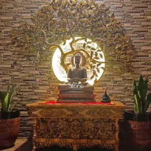 beautifully curated and serene display, likely showcasing premium products from "The Stone Studio," is the focal point against a textured wall made of stacked natural stone veneer in varying warm tones of beige, tan, and light brown. Centrally positioned is a dark stone Buddha statue, sculpted in a contemplative meditative posture (Dhyana Mudra), seated upon a multi-tiered lotus pedestal. Behind the Buddha, a large, circular back-lit panel emits a soft, inviting glow, creating a radiant halo effect. Ornate golden metallic branches and delicately sculpted leaves, resembling a stylized Bodhi tree or Tree of Life, gracefully fan out from this illuminated circle, suggesting spiritual enlightenment and natural harmony. The Buddha statue rests on an intricately carved wooden altar or console table made from rich, dark wood, showcasing traditional craftsmanship with elaborate patterns that include what appear to be dragons, floral motifs, and possibly Dharma wheels. A vibrant red fabric runner drapes across the tabletop, adding a striking contrast of color. To the right of the Buddha, a small, black ceramic incense burner is visible on the table, contributing to the tranquil atmosphere. Flanking the wooden altar on both sides are tall, vibrant green snake plants (Sansevieria) housed in decorative, textured terracotta pots, which add a touch of natural vitality and balance to the composition. The overall lighting is warm and ambient, highlighting the central display and creating a peaceful, Zen-like ambiance. The "the Stone STUDIO" logo is subtly overlaid on the lower body of the Buddha, emphasizing the image's connection to high-quality stone artifacts, interior decor, spiritual home accents, and bespoke stone and wood craftsmanship. This image powerfully conveys themes of tranquility, spiritual decor, premium home design, and the beauty of natural materials.
