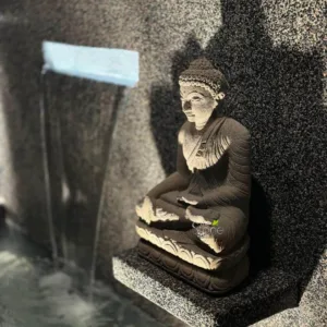 serene and contemplative scene features a meticulously carved stone Buddha statue positioned beside a modern water feature, set against a richly textured wall. The Buddha, crafted from light-hued, possibly sandstone or cast stone, sits gracefully in a meditative Dhyana Mudra pose, with hands resting in its lap and legs crossed. Its serene expression, downcast eyes, and finely detailed features, including the traditional ushnisha and intricate folds of its draped robes, convey a profound sense of peace. The statue rests on a beautifully sculpted lotus pedestal, which in turn is placed upon a polished, dark gray granite-like shelf that extends from the wall. To the left, a sleek, rectangular light-colored spout is integrated into the textured wall, from which a thin, continuous sheet of water gently cascades downward. The flowing water creates a soothing visual and auditory element, adding to the tranquil ambiance as it falls into a dimly visible, rippling pool below. The entire setting is framed by a striking wall clad in dark, speckled natural stone, possibly polished granite or basalt, whose coarse texture is dramatically highlighted by directional lighting from above and to the right. This light casts soft shadows that emphasize the depth and detail of both the stone wall cladding and the statue. The interplay of light and shadow, the rough texture of the natural stone contrasting with the smoother, sculpted Buddha, and the dynamic flow of water create a harmonious and inviting atmosphere, perfect for a Zen garden, spa, or meditation space. This image beautifully illustrates the quality and versatility of stone products in creating exceptionally peaceful and aesthetically pleasing environments.