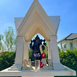 beautifully crafted, dark-colored statue of the Hindu deity Lord Ganesha stands prominently within a modern, light-toned stone shrine on a bright, sunny day, surrounded by lush greenery. The Ganesha statue, appearing to be made of black or very dark grey polished stone, features his iconic elephant head, four arms, and rotund belly, symbolizing prosperity and wisdom. He is adorned with a thick, fragrant-looking white flower garland draped elegantly around his neck and shoulders, and a smaller red and white floral garland near his feet. A subtle red tilak (mark) adorns his forehead. The shrine itself is a striking example of contemporary design, constructed from a light, speckled stone or terrazzo material that gives it a pristine, clean appearance. Its roof is distinctively layered and stepped, forming an angular, inverted pyramid shape that creates a protective canopy over the deity. This structure is supported by four sturdy, square pillars, which frame Ganesha centrally. The shrine sits on a robust, multi-tiered base which serves as an altar, meticulously designed with clean lines and sharp edges, showcasing a blend of traditional reverence with modern architectural aesthetics. On this altar, several devotional items are thoughtfully arranged: two small, elegant white elephant figurines with subtle gold accents flank the statue, facing inwards, symbolizing auspiciousness. A slender white horse figurine stands near the center, alongside a clear glass tumbler and smaller floral offerings in shades of red and white. The entire outdoor setting is vibrant and inviting; to the left, lush green trees with delicate, weeping willow-like branches are visible against a brilliant, cloudless blue sky, adding a natural, serene backdrop. To the right, a portion of a modern, light-colored house (likely white or cream) with a dark tiled roof can be seen, partially obscured by neatly trimmed green bushes, suggesting a private garden, home, or residential compound. This image effectively captures the spiritual tranquility and artistic integration of a meticulously designed personal or home shrine, emphasizing both the devotional artistry and the modern architectural use of stone in creating sacred spaces.