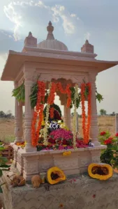 beautifully detailed image captures a newly installed, ornate stone shrine or miniature temple, likely a custom-built mandap, set outdoors under a bright blue sky dotted with soft white clouds. The structure, crafted from a pale, creamy beige or light pink sandstone, features intricate carvings on its four supporting pillars and base, showcasing delicate floral and geometric patterns. A rounded dome (shikara) caps the roof, flanked by smaller decorative finials, highlighting traditional Hindu architectural elements. The shrine is lavishly adorned for a ceremonial occasion, such as a pooja or land dedication (Bhumi Pujan). Long, vibrant garlands of orange marigold-like flowers and contrasting white jasmine or tuberose flowers, interspersed with fresh green leaves, drape gracefully from the roof. Inside the shrine, a central idol or deity, mostly obscured, is covered with a bountiful mound of fresh flower petals in shades of pink, red, yellow, and white, suggesting deep devotion. On the rough concrete platform elevating the stone shrine, traditional offerings are carefully arranged: two whole coconuts (one broken open to reveal its white flesh), a small metal container, and two large, halved pumpkins or squash displaying their bright orange interior and seeds. Scattered flower petals are also visible on the concrete surface, adding to the ceremonial atmosphere. In the background, a few red flowering plants add a touch of color. The setting is an expansive, dry, yellowish-brown field, possibly agricultural land or an undeveloped rural area, stretching towards a distant horizon with sparse trees under the clear sky. A subtle watermark or logo for "The Stone Studio" is visible over the flower offerings inside the shrine, indicating the craftsmanship and source of this custom stone temple. The image effectively conveys a scene of spiritual significance, celebrating a new beginning through traditional ceremony and expert stone masonry.