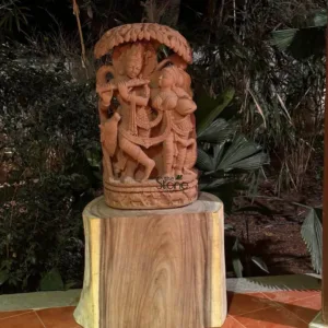 long and descriptive alt text for this image: A beautiful, hand-carved reddish-brown stone sculpture depicting the divine Hindu deities Lord Krishna and Radha, displayed prominently outdoors or in a garden setting at night. On the left, Lord Krishna is intricately carved, shown playing his characteristic flute, standing gracefully with one leg bent at the knee, wearing a detailed crown and traditional dhoti-like garments. A small calf or cow is sculpted near his leg, symbolizing his role as a cowherd and echoing the pastoral setting of Vrindavan. To his right, Radha stands elegantly, holding an offering pot or kalash, her gaze directed towards Krishna, her spiritual beloved. She too is adorned with intricate jewelry and a crown, her traditional attire complementing Krishna's. Above the figures, a finely sculpted tree canopy, possibly representing a Kadamba tree with its distinctive leaves, arches protectively, adding to the naturalistic and romantic scene. The entire ensemble rests on a multi-tiered circular base, which is also intricately carved with smaller animal figures and foliage, further enhancing the pastoral narrative. This masterpiece of traditional Indian art is displayed atop a rustic, substantial wooden pedestal or tree stump, showcasing its natural wood grain and providing a sturdy, organic foundation. The setting is dimly lit, likely at night or in a shaded area, with lush, dark green foliage and palm-like plants forming a soft, blurred background, which enhances the spiritual and serene ambiance of the sculpture. The warm, earthy tones of the stone and the natural wood contrast gently with the deep greens and shadows of the surroundings, making the devotional sculpture a captivating focal point. This detailed stone carving, rich in Hindu mythology and exemplary Indian craftsmanship, serves as a significant piece of spiritual art, ideal for garden decor, temple settings, or cultural collections, embodying themes of love, devotion, and nature.