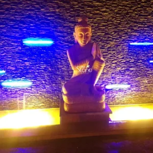 "A beautiful and serene Buddha statue, carved from light-toned, earthy-colored stone, is centrally positioned in a dimly lit, dramatic setting. The Buddha is depicted in a meditative, seated pose, likely Padmasana (lotus position), with both hands raised and clasped together in the Anjali Mudra (prayer or greeting gesture) at chest level. The statue's face emanates tranquility with gently downcast eyes and a subtle, peaceful smile. Distinct traditional features such as the ushnisha (a protuberance on the crown of the head symbolizing spiritual enlightenment) and elongated earlobes are clearly visible. The Buddha rests upon a multi-tiered, intricately carved stone pedestal, which adds to its grandeur. The background features a dark, textured wall, appearing to be constructed from irregularly stacked, rough-hewn natural stone or split-face stone tiles, creating a rugged yet sophisticated surface. Embedded horizontally across this wall are several vibrant, cool blue LED-like light strips, casting an ethereal, modern glow that contrasts sharply with the dark stone texture. From below the Buddha statue, a powerful warm golden-yellow light emanates, illuminating the base of the statue and casting a soft, inviting radiance upwards onto the Buddha's lower body and the bottom portion of the textured wall. This interplay of cool blue and warm yellow light creates a striking visual depth and a contemplative ambiance. The entire scene is enveloped in darkness, allowing the illuminated stone elements to stand out prominently. A subtle 'The Stone Studio' watermark is visible on the Buddha's chest, indicating this exquisite piece showcases premium stone craftsmanship and innovative design. This image effectively highlights a sophisticated blend of spiritual tranquility and contemporary aesthetic, suggesting its use in high-end home decor, serene garden landscapes, or as a focal point in architectural design that leverages custom stone sculptures and strategic mood lighting to create a peaceful and luxurious atmosphere."